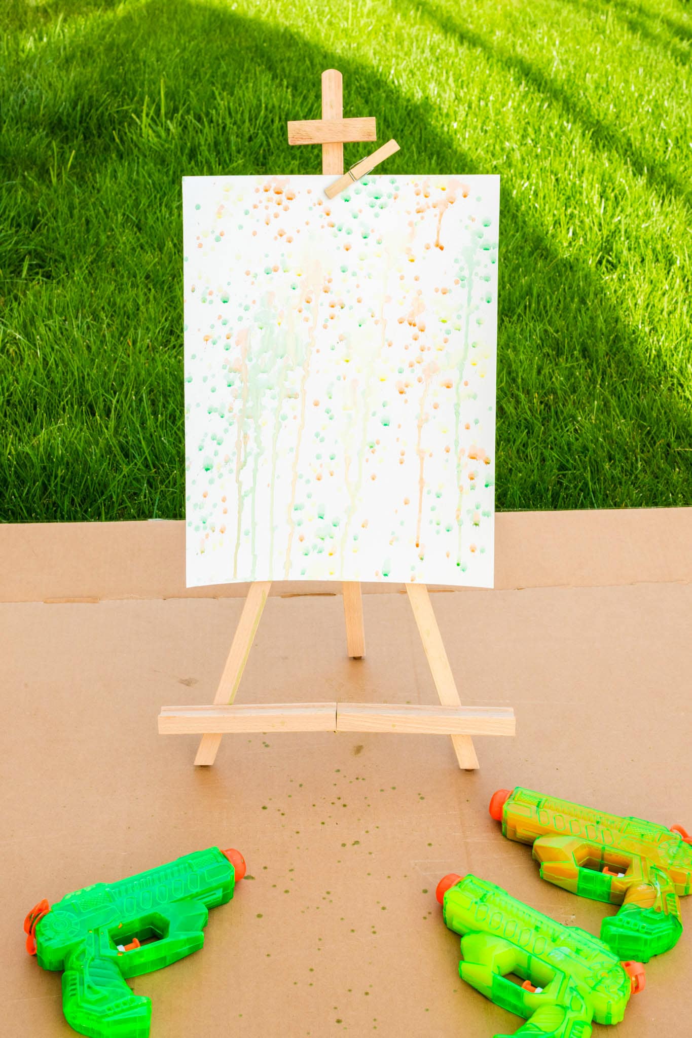 A nearly completed splatter painting on a wooden easel outdoors, showing dense patterns of green, orange, and yellow paint dots and drips, with neon water guns placed on the ground in front.