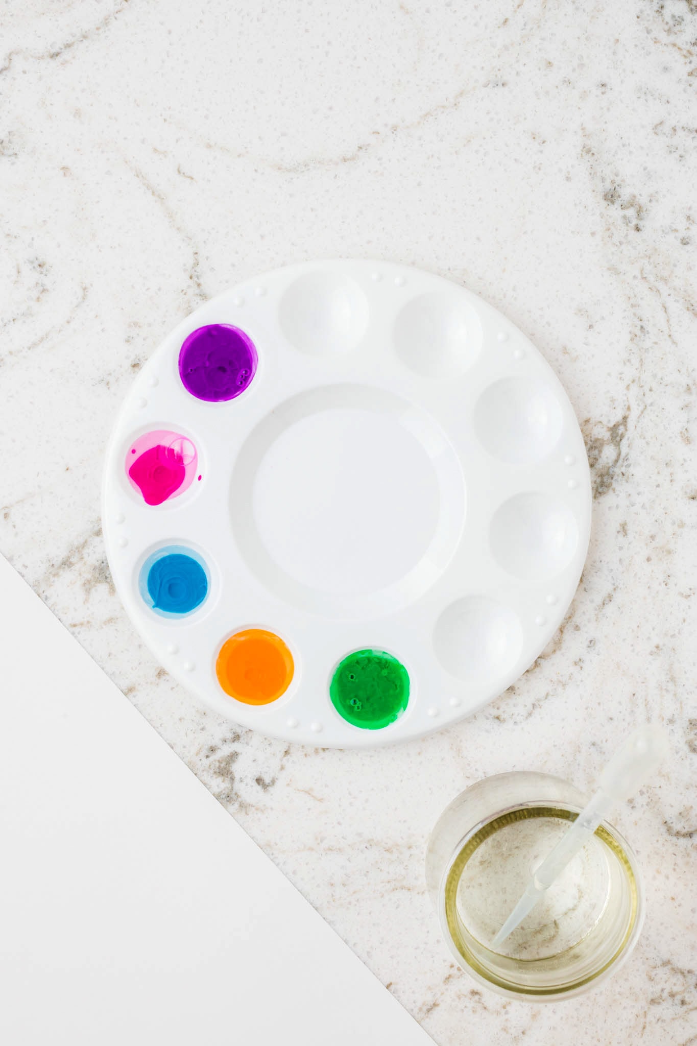 A plastic paint palette containing five wells filled with bright liquid watercolors (purple, pink, blue, orange, and green), placed next to a small jar of water with a plastic dropper.