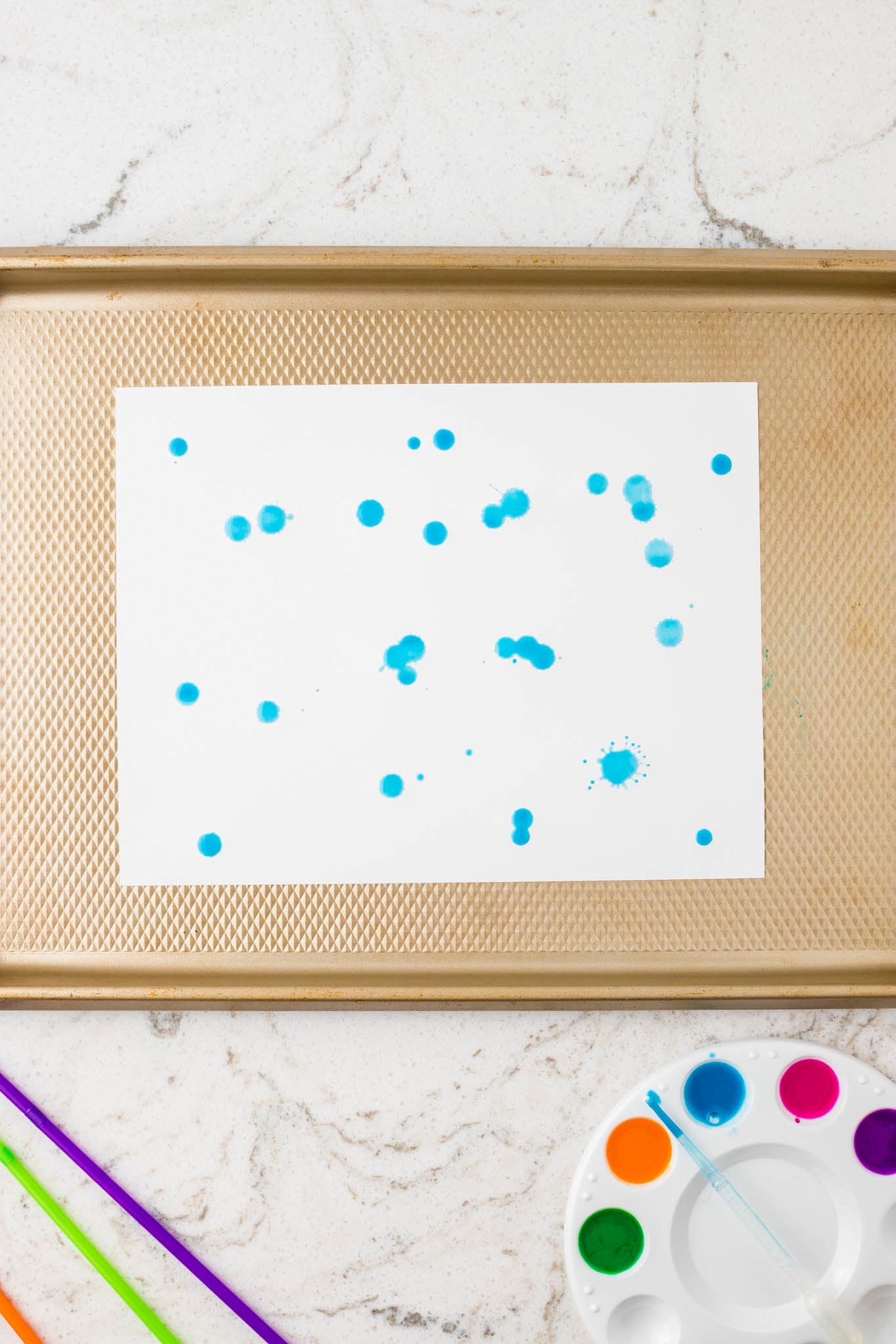 A piece of white paper with scattered drops of blue paint, placed on a gold baking tray. Colorful straws and a paint palette are visible nearby.