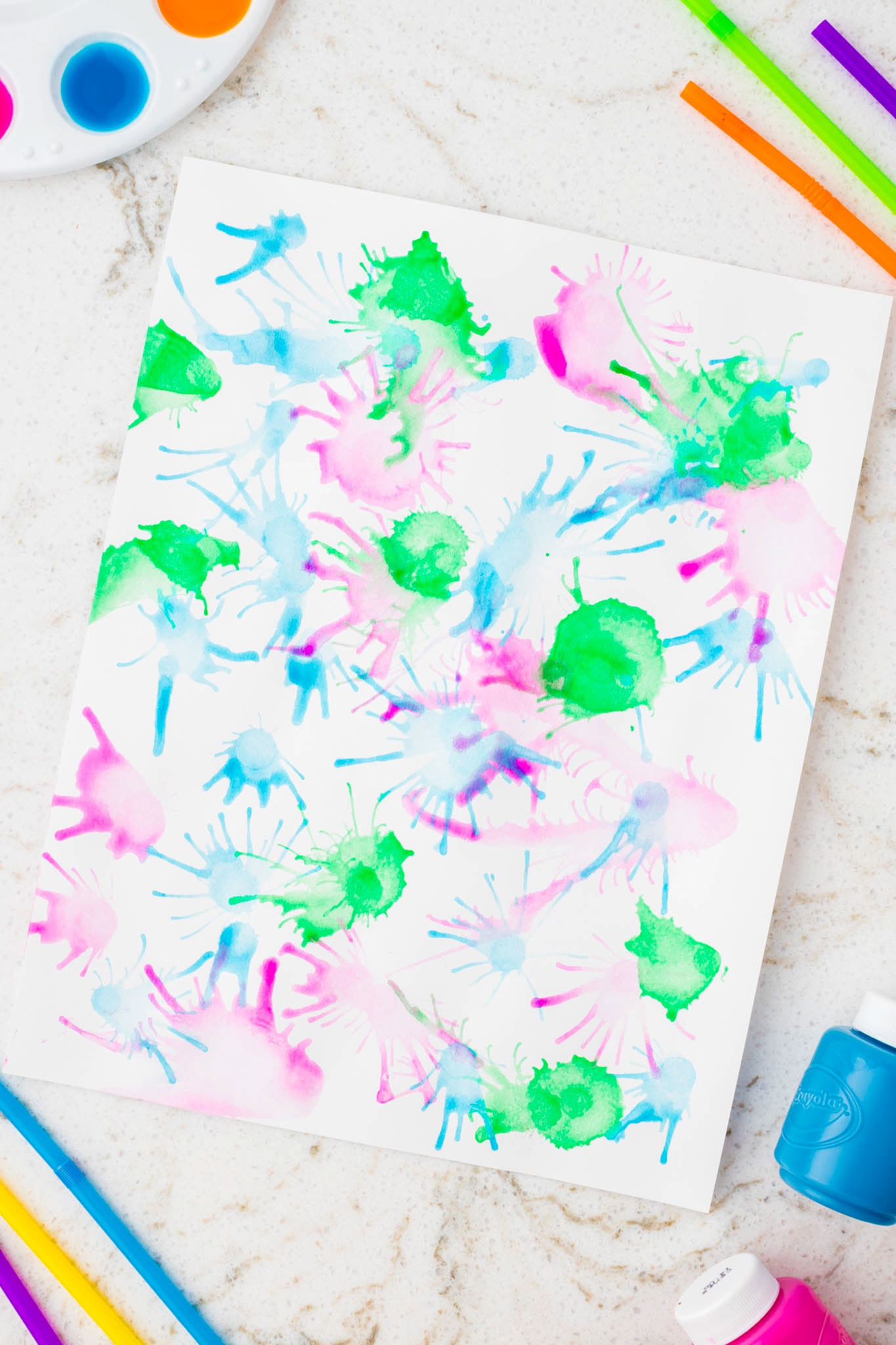 A closer view of a colorful blow painting with pink, green, and blue radial paint bursts spread across the white paper.