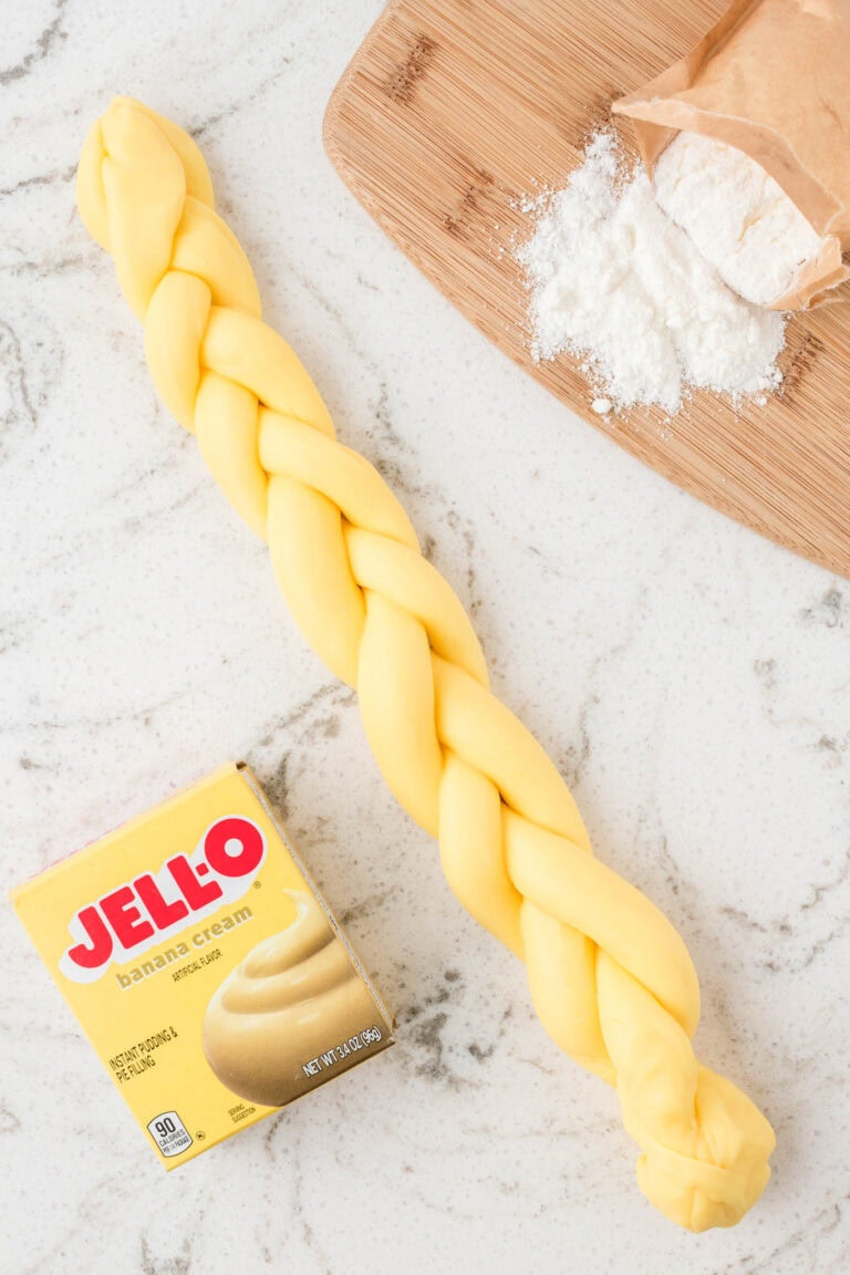 A long braid of smooth, bright yellow pudding slime stretched out on a countertop next to the pudding mix box and some spilled white powder, showing the final fun texture.