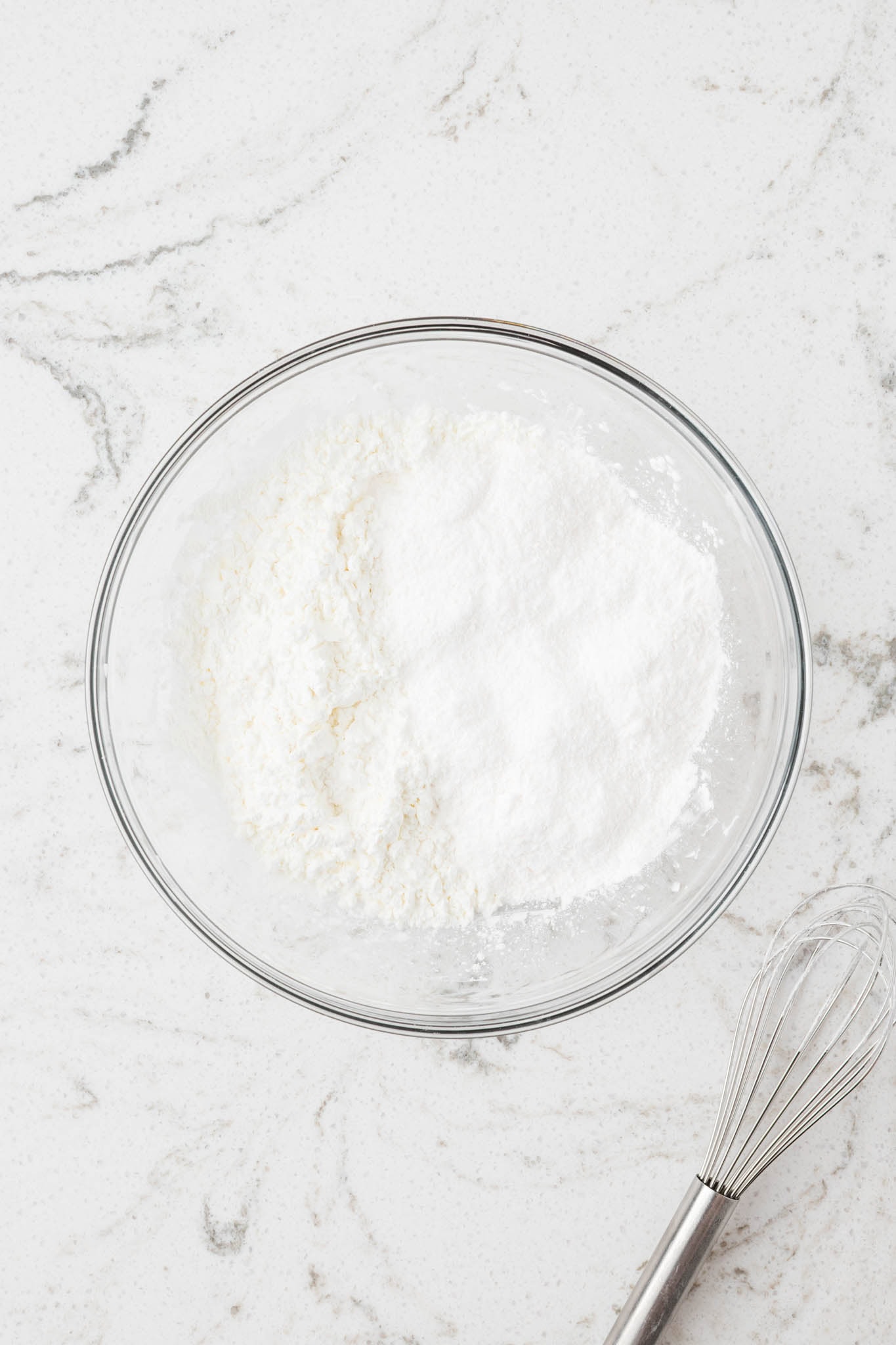 A glass mixing bowl filled with a white powder mixture, likely cornstarch or pudding mix, sitting on a white marble countertop next to a whisk.