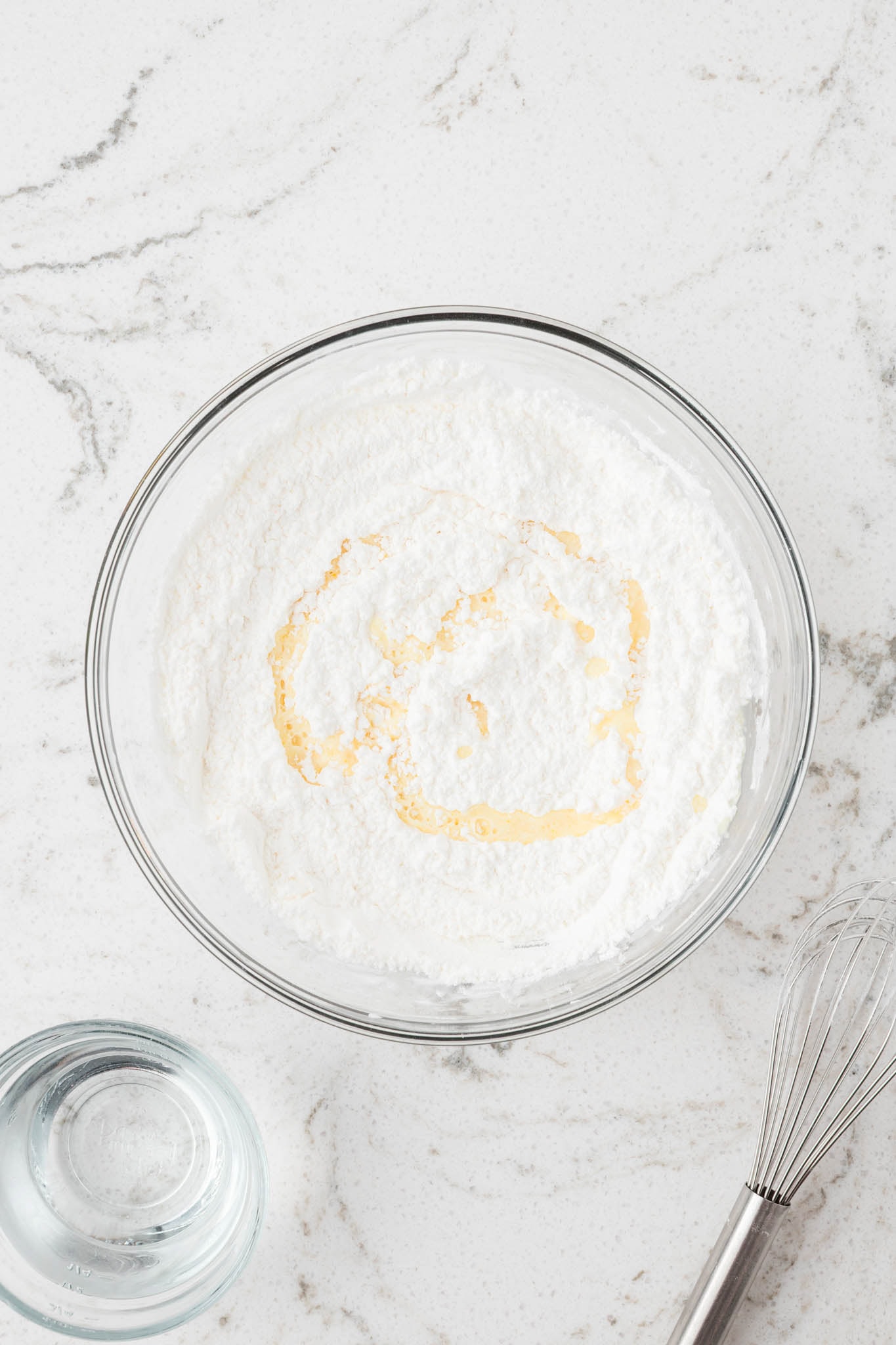 The same bowl of white powder now with a swirl of yellow-tinted liquid being added, likely vanilla extract or a similar ingredient.