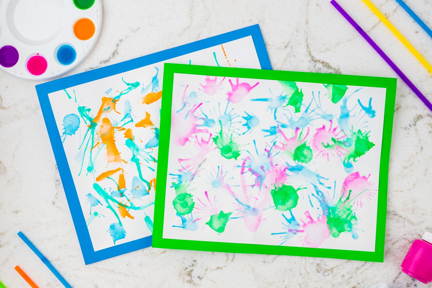 Two abstract paintings created using the straw blow painting technique. Each sheet is framed with a colored border (blue and green), featuring splatters and streaks of vibrant watercolor paint in colors like green, orange, blue, and pink.