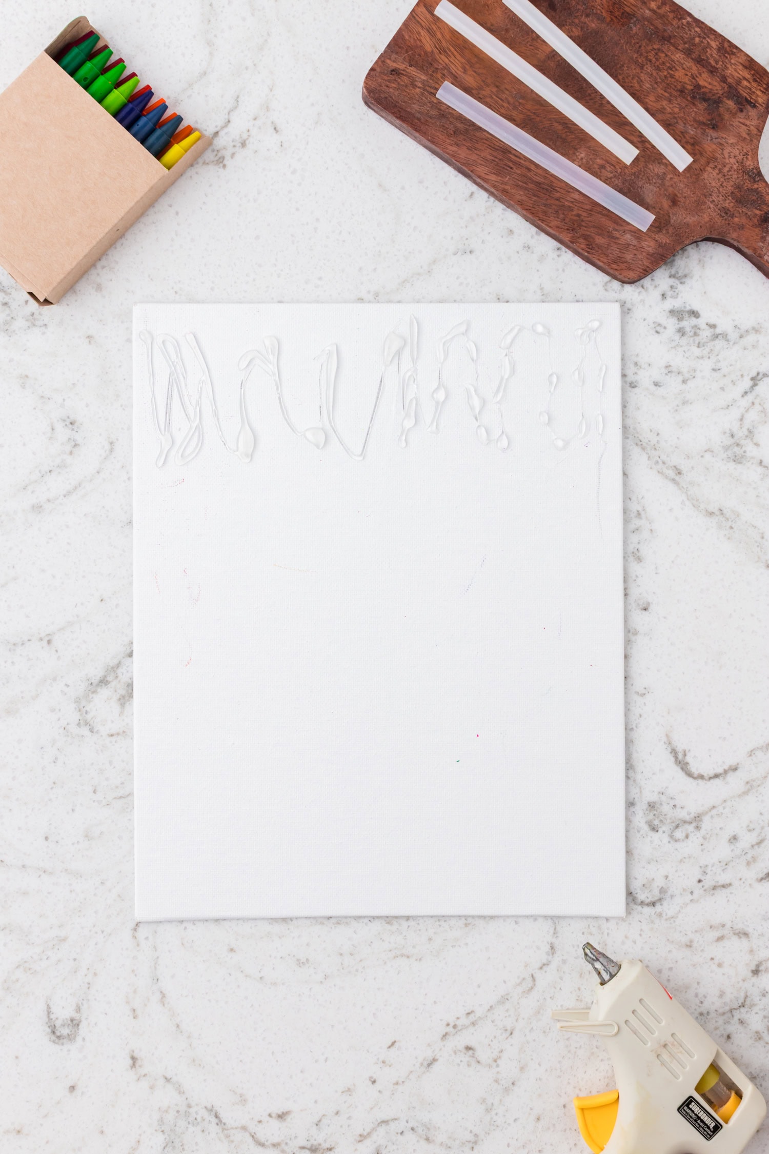 A blank white canvas with hot glue squiggles applied along the top edge. A glue gun, glue sticks, and a box of crayons are nearby on a marble surface.
