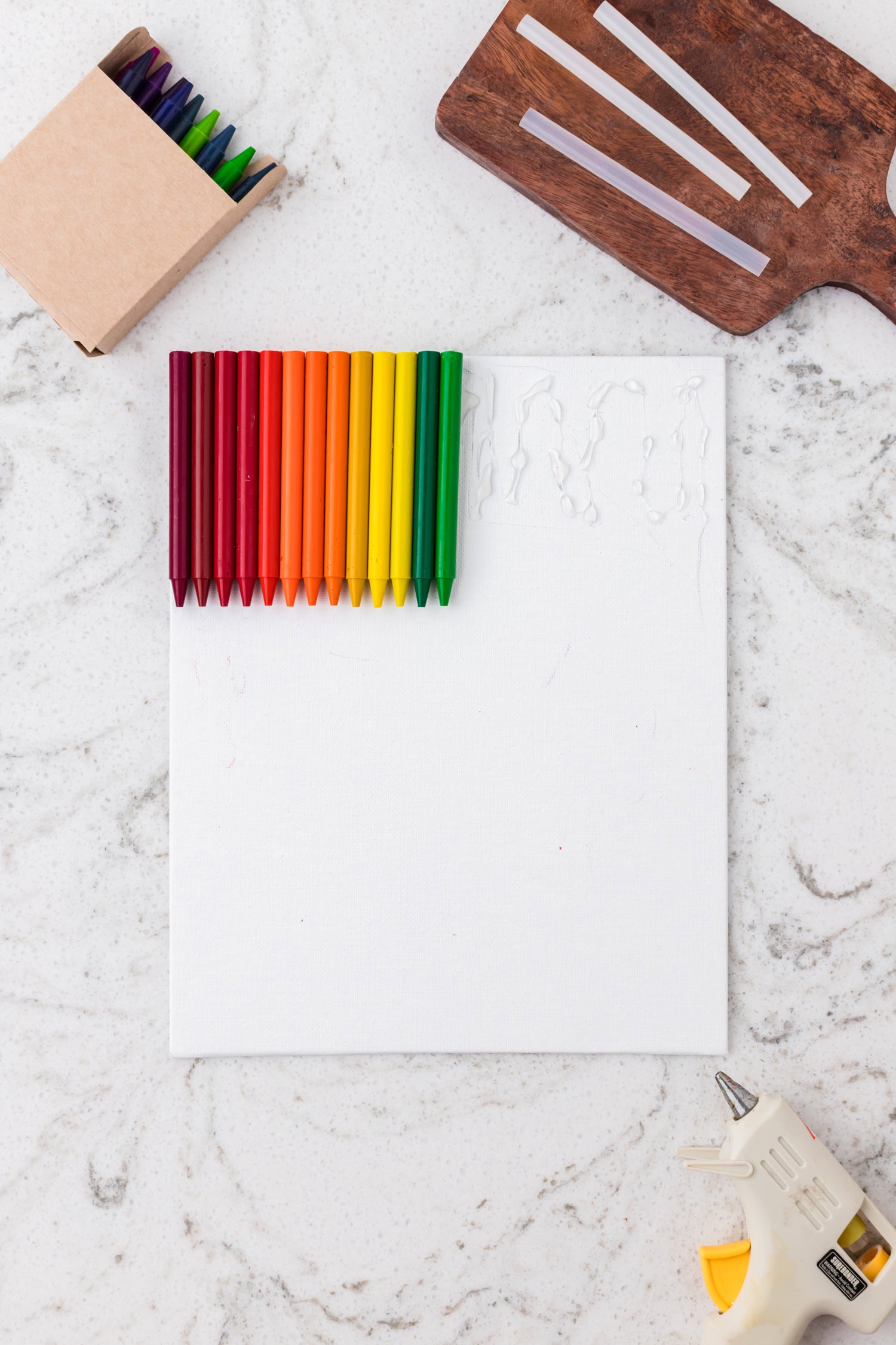 Several crayons in red, orange, yellow, and green shades have been glued to the top of the canvas in a row, over the dried glue.