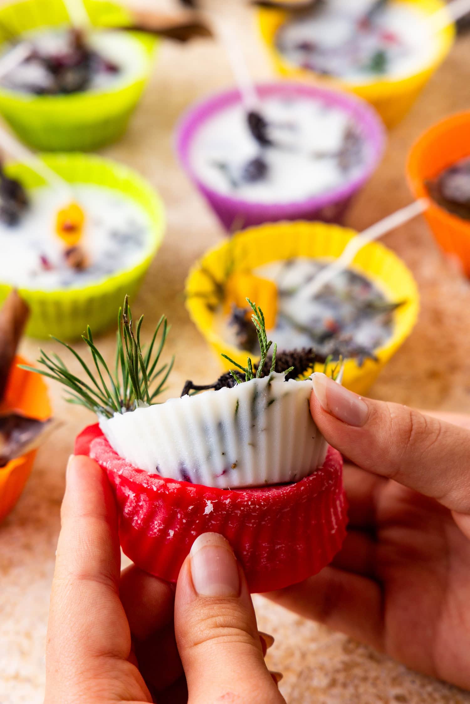 A person’s hands peel back the red silicone liner to reveal a finished campfire starter with embedded herbs and a centered wick.