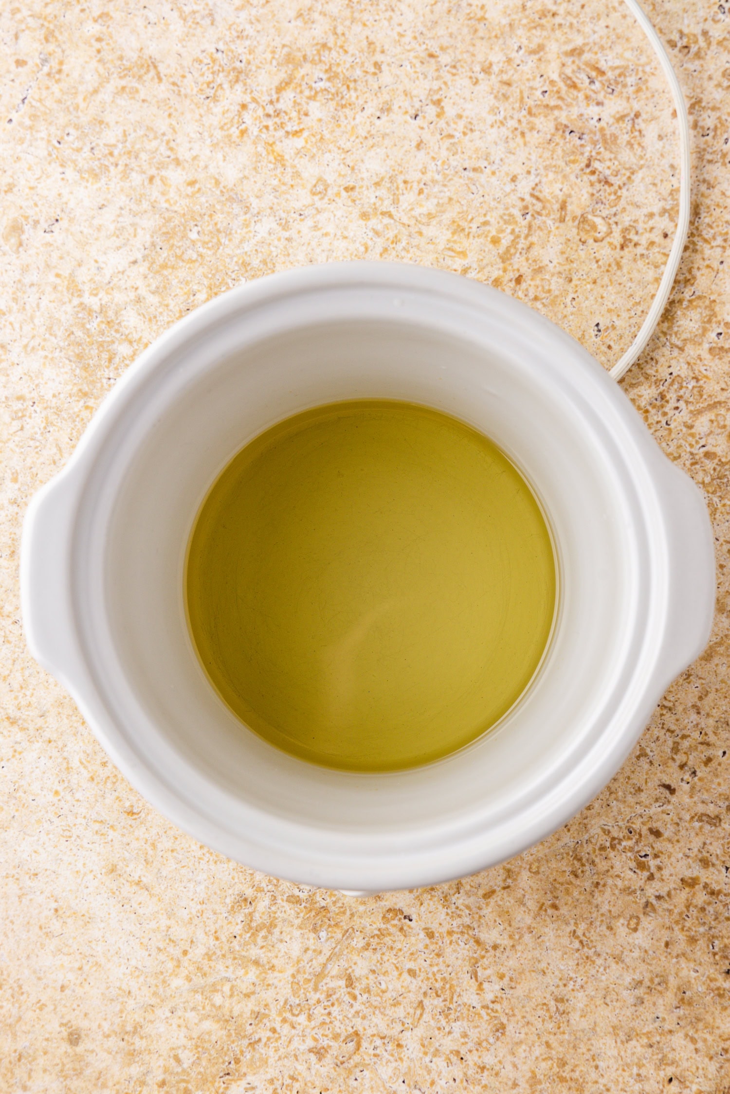 A white ceramic crockpot with melted wax inside, sitting on a beige marbled countertop. The wax is translucent and yellowish in color.
