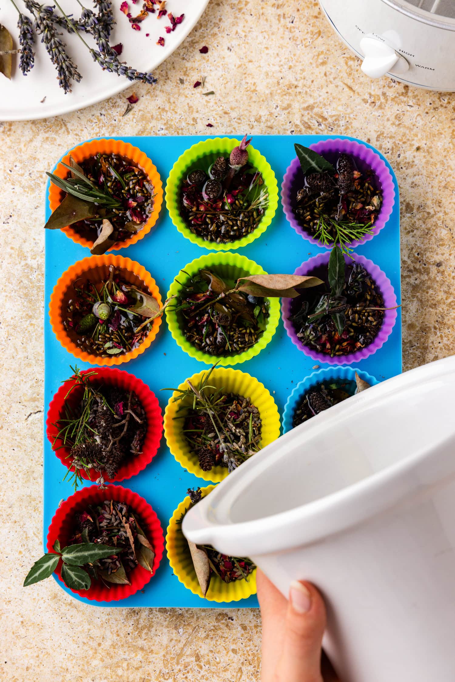 The melted wax is being poured from the white crockpot into the colorful silicone liners containing dried botanicals.