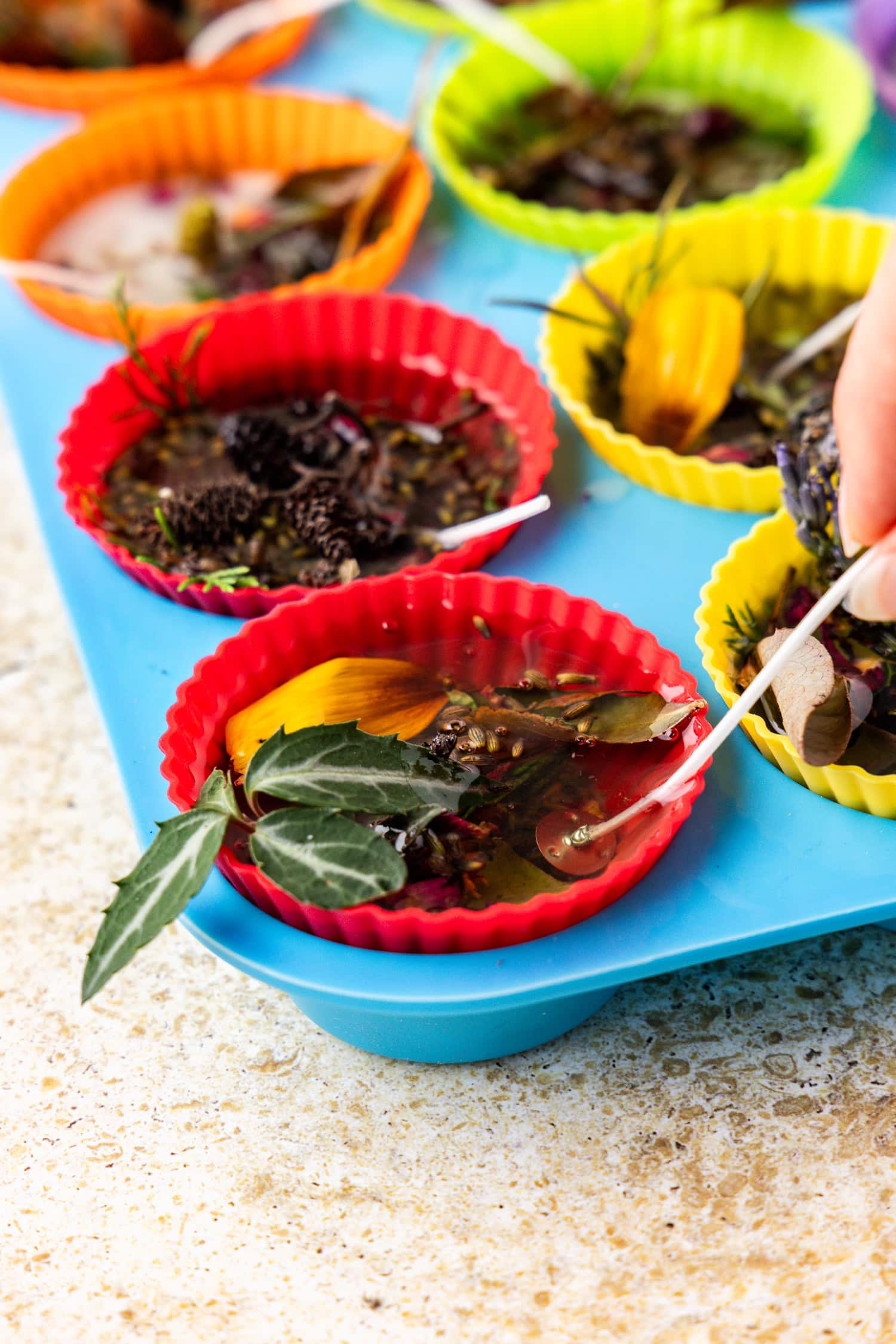 A close-up of the silicone liners now filled with wax and botanical mixtures. A hand is inserting a wick into one of the wax-filled molds.