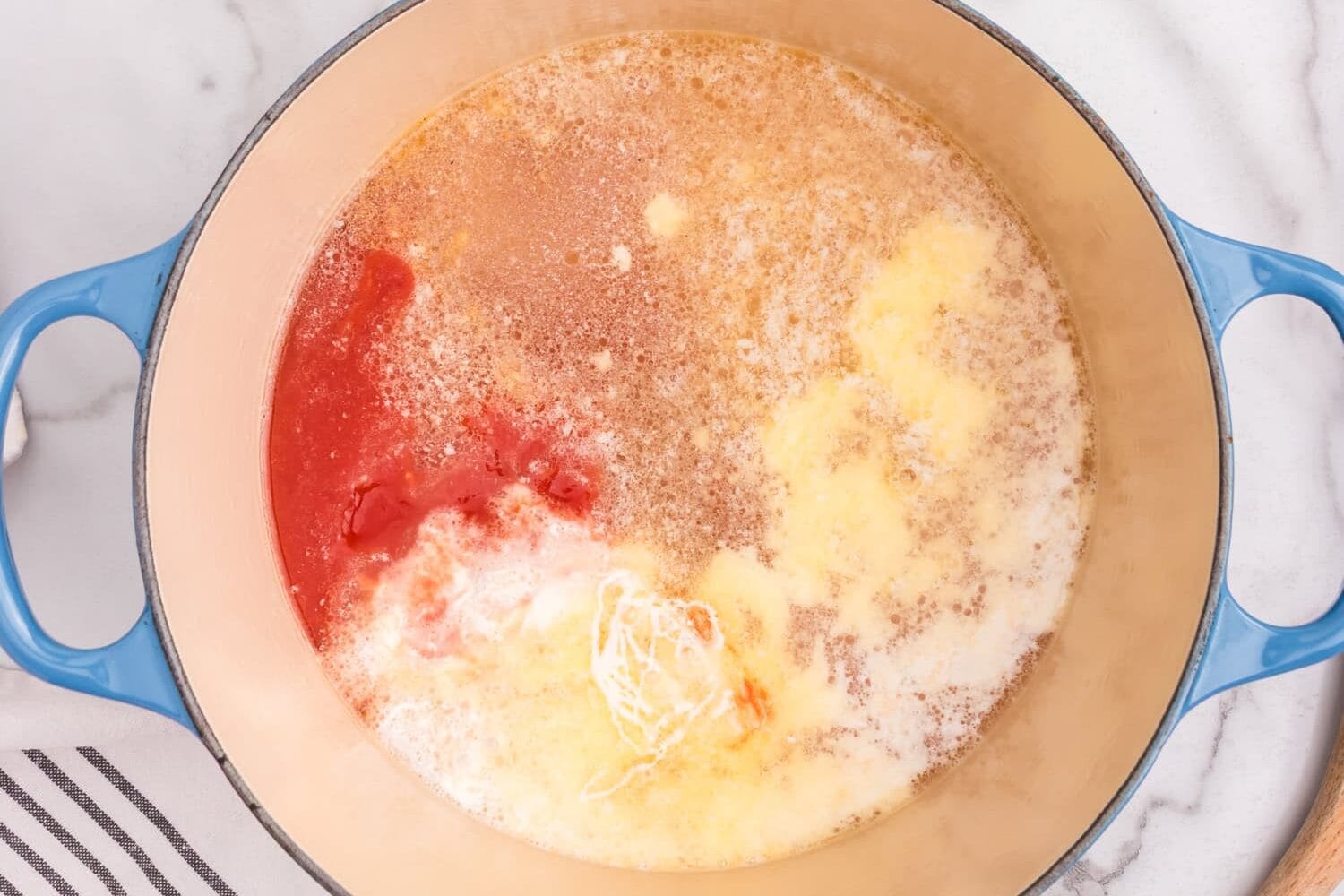 The Dutch oven filled with melted butter, tomato sauce, milk, and seasonings before being stirred together. A bowl of dry pasta rings and shredded cheese are visible nearby.