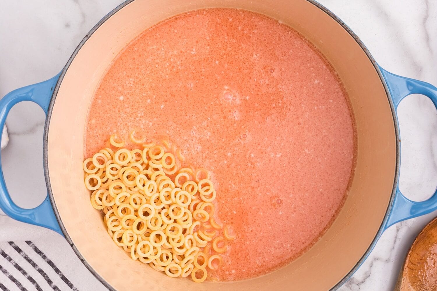 The tomato and cream mixture in the pot, with uncooked pasta rings added in. A bowl of shredded cheese sits beside the pot.