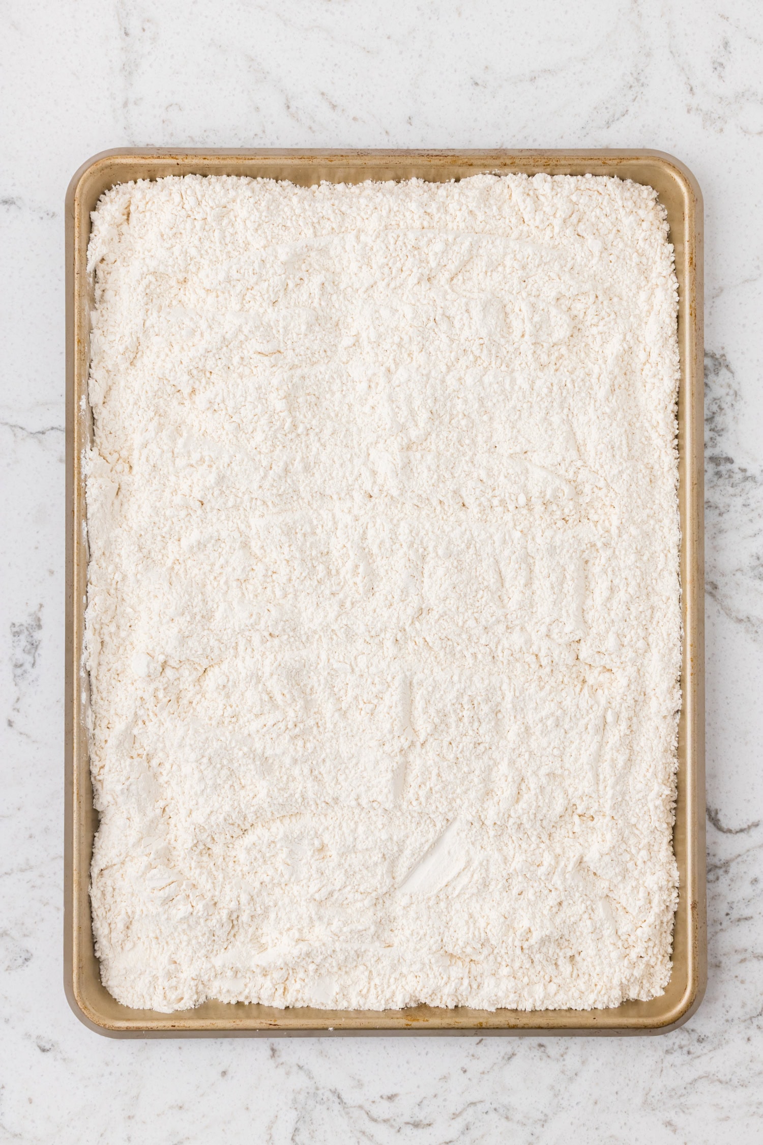 A baking sheet filled with a layer of flour spread evenly across the surface on a marble countertop.