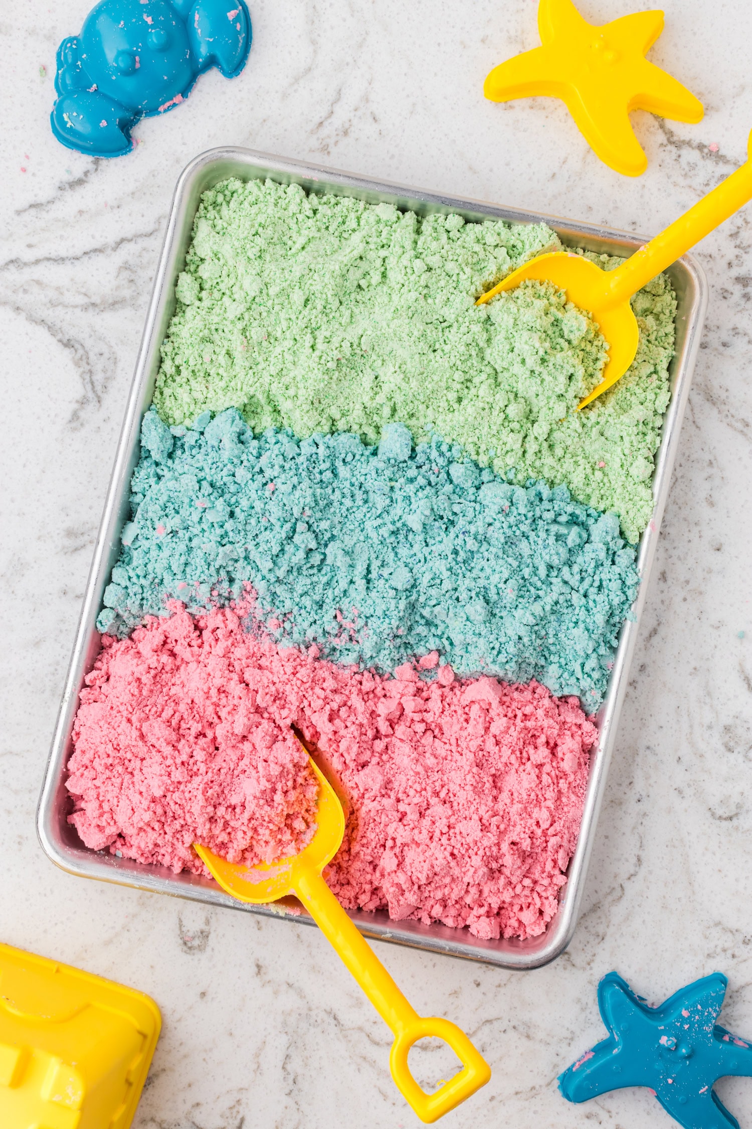 A tray filled with green, blue, and pink moon sand in neat rows, with yellow plastic toy shovels placed inside.