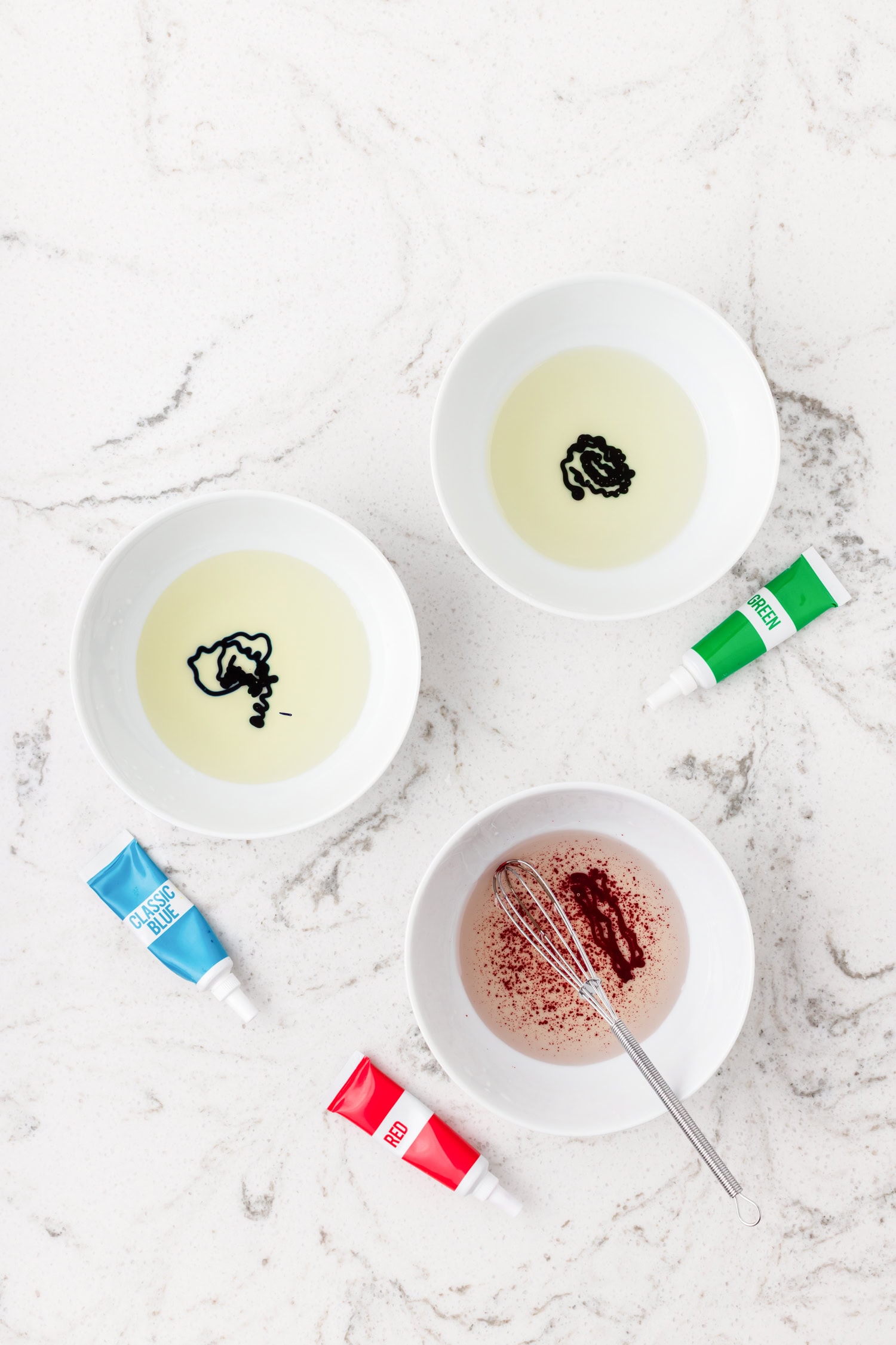 Three bowls of oil each with food coloring drops (blue, green, red) on a marble surface with food coloring tubes beside them.