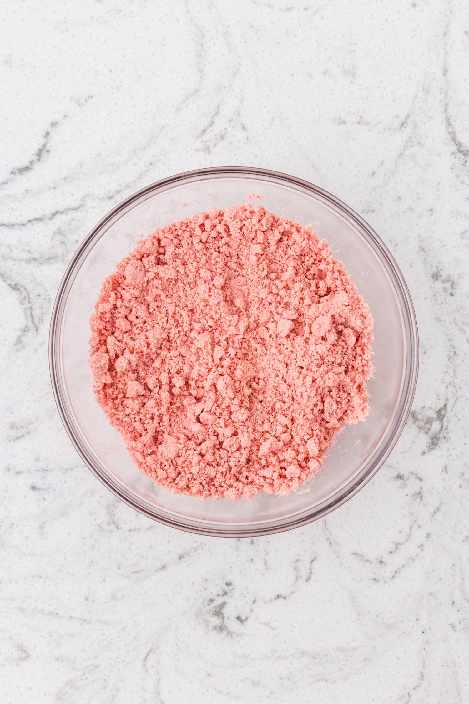 A glass mixing bowl of flour mixture evenly blended into a crumbly pink sand-like texture.