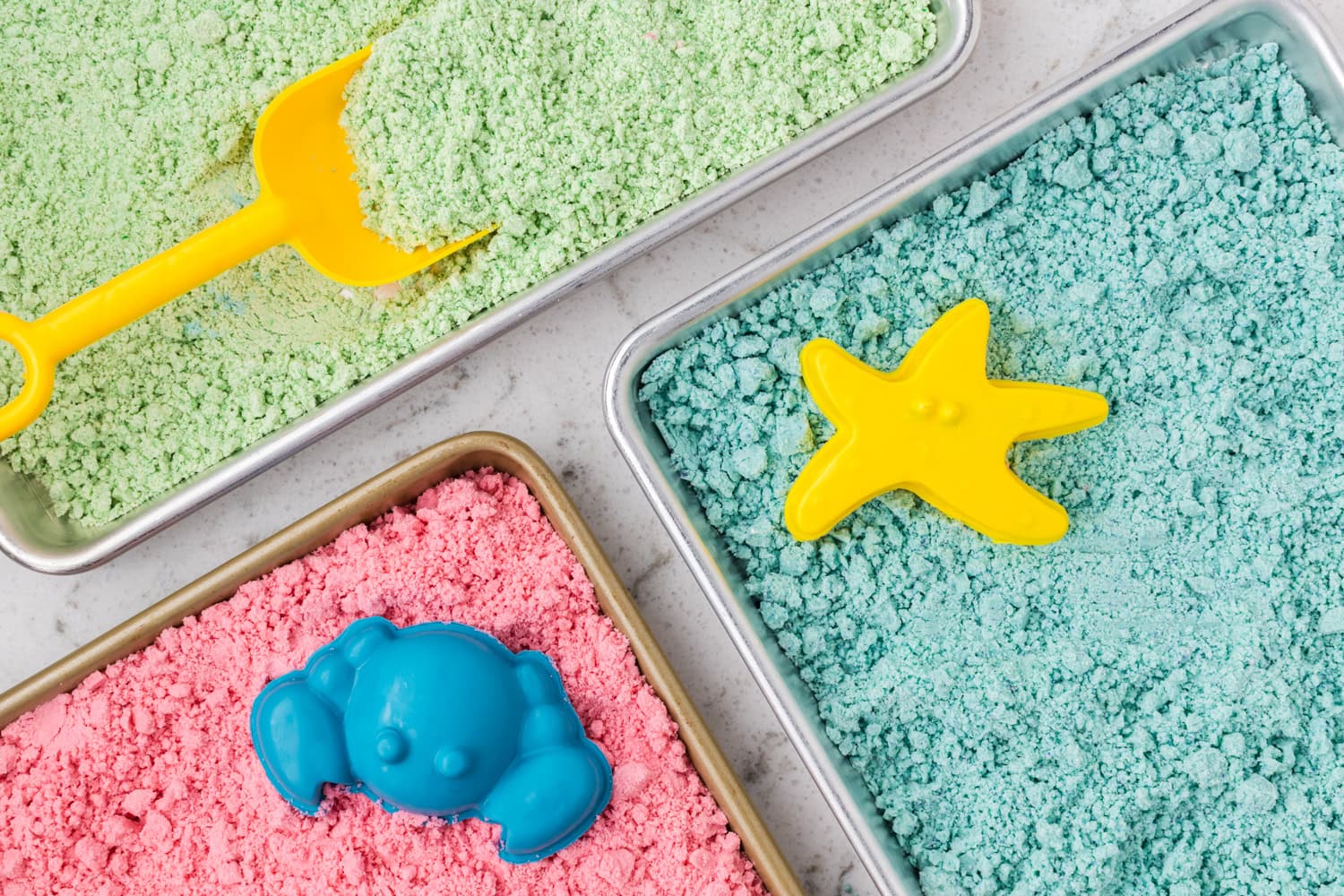 Three trays filled with pink, blue, and green moon sand, each with yellow or blue plastic molds shaped like a crab and starfish.