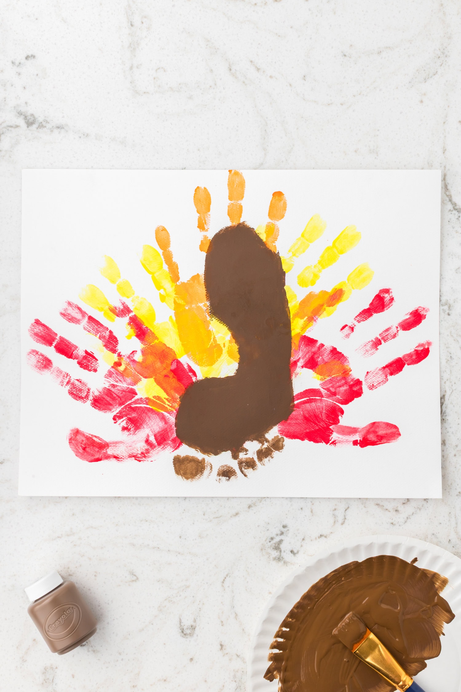 A painted turkey craft in progress — colorful handprint feathers in the background and a brown footprint added on top to form the turkey’s body.
