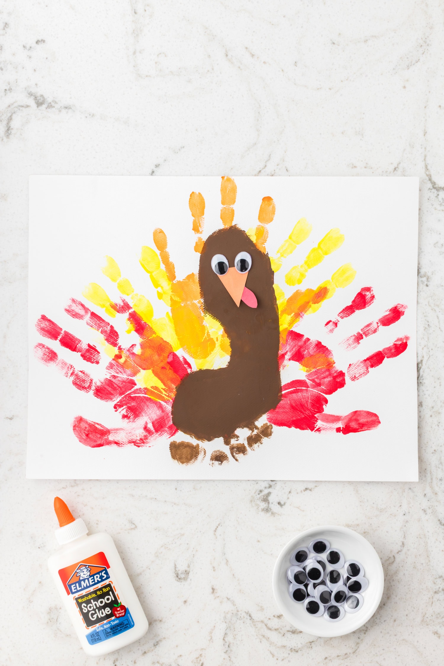 The nearly finished turkey craft featuring colorful handprint feathers, a brown footprint body, and added googly eyes, beak, and wattle; glue and googly eyes are visible on the table.
