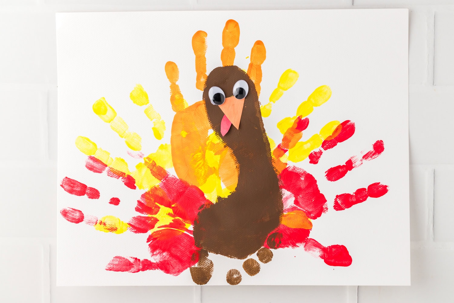A completed Thanksgiving turkey craft made with colorful handprints in red, yellow, and orange for feathers, a brown footprint for the body, and added googly eyes, an orange beak, and a red wattle on white paper.