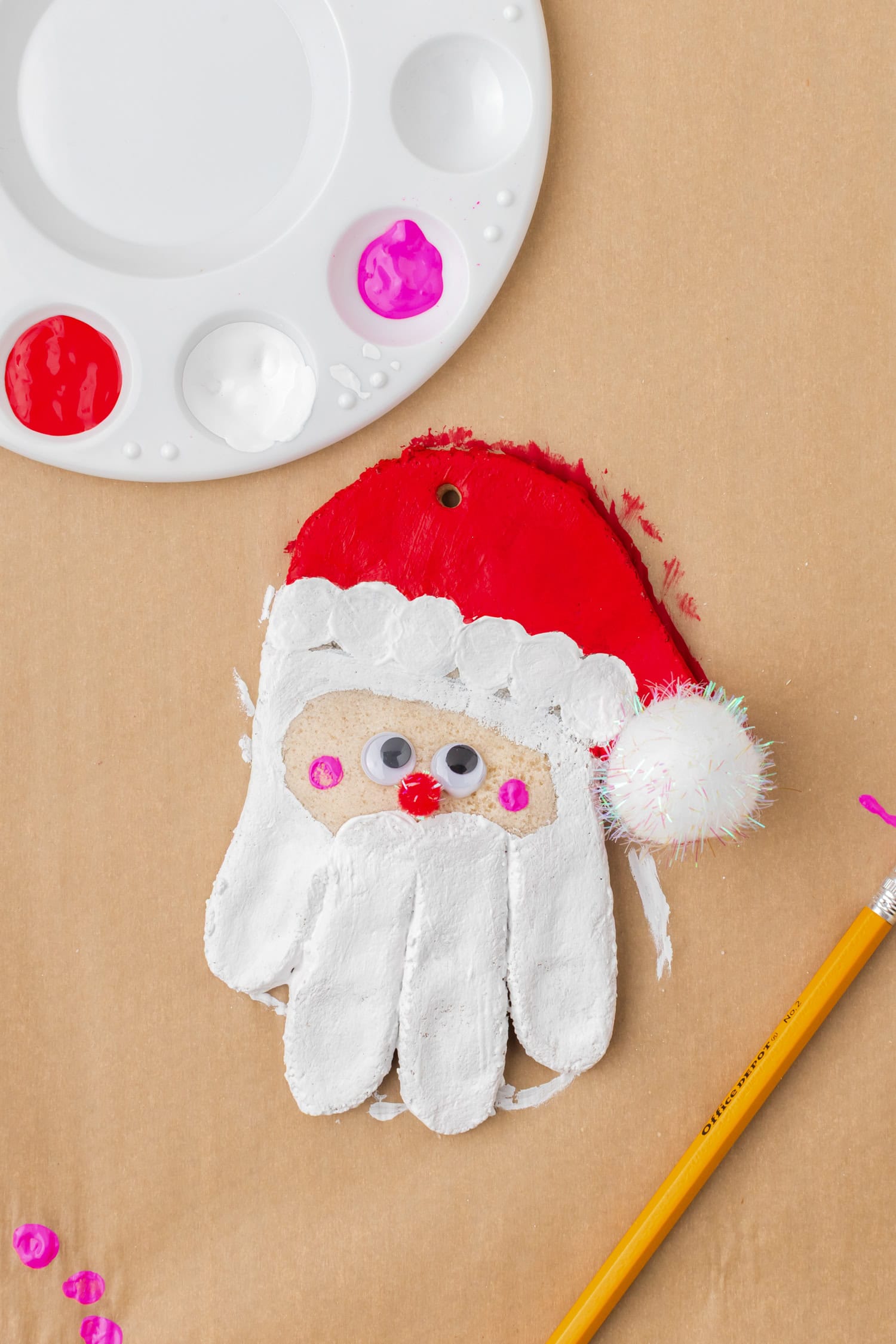 Partially painted handprint ornament decorated as Santa Claus with red, white, and pink paint, googly eyes, and pom-pom hat.