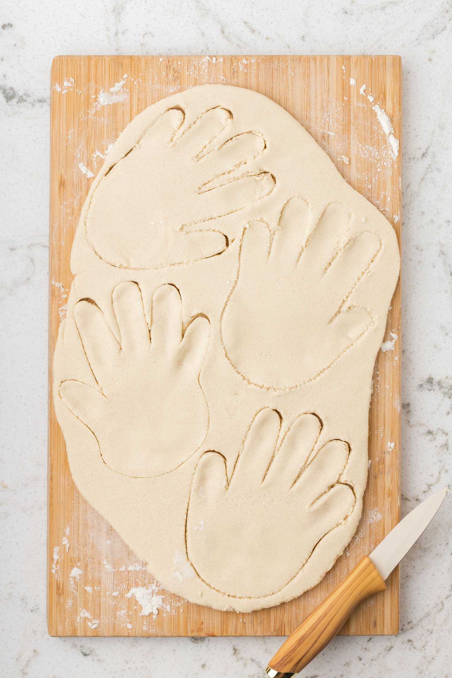 Rolled-out salt dough on a cutting board with handprint shapes cut out using a knife.