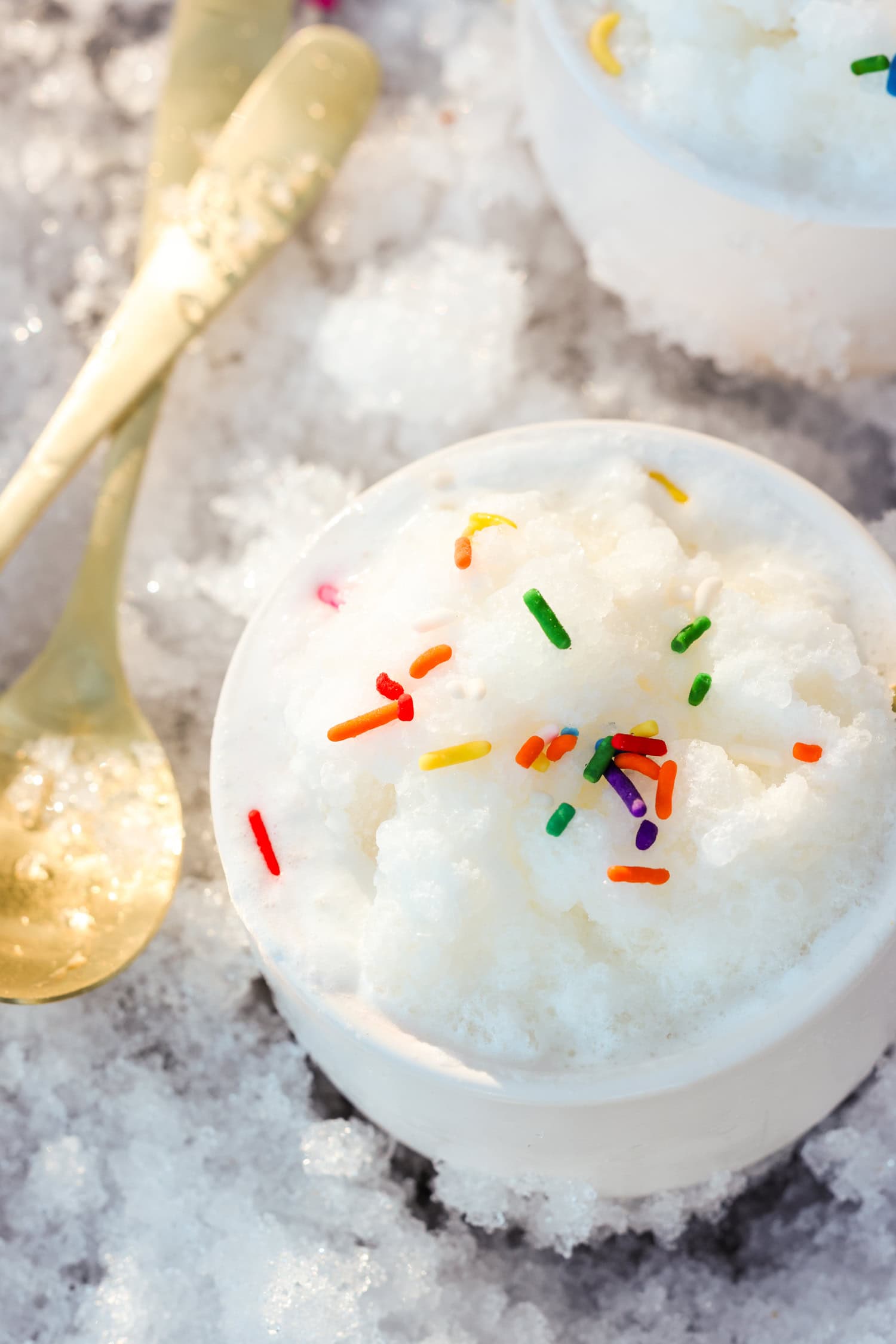 A bowl of fluffy snow cream topped with colorful sprinkles, placed on a snowy surface, with a golden spoon resting nearby.
