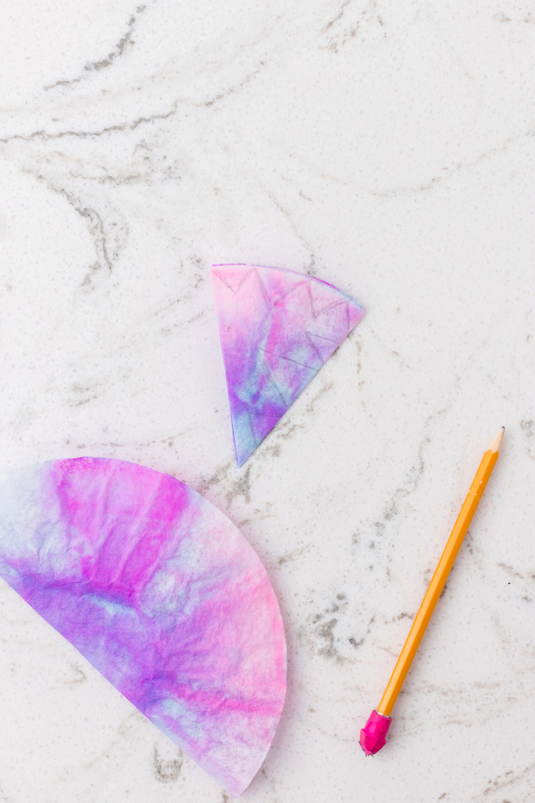 Folded and colored coffee filter in shades of pink and purple on a white marble surface, with a pencil showing snowflake cutting guides.