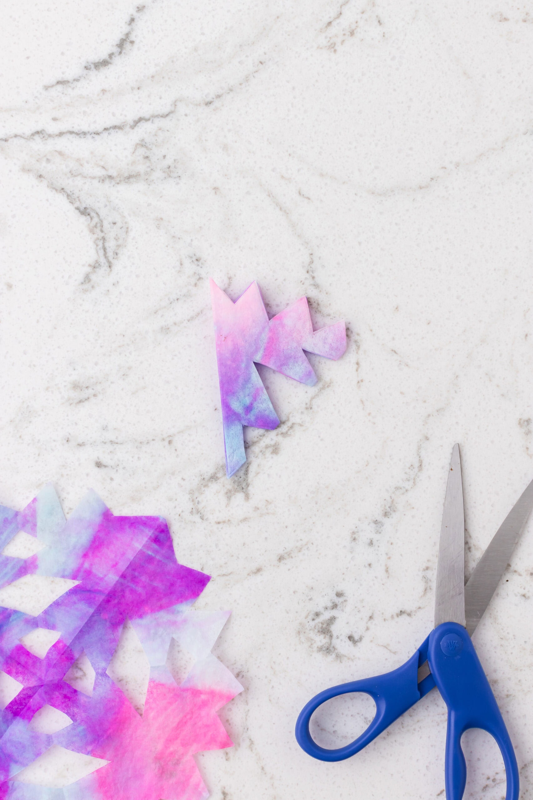 Folded, dyed coffee filter with cut-out snowflake shapes next to blue scissors on a white marble surface.