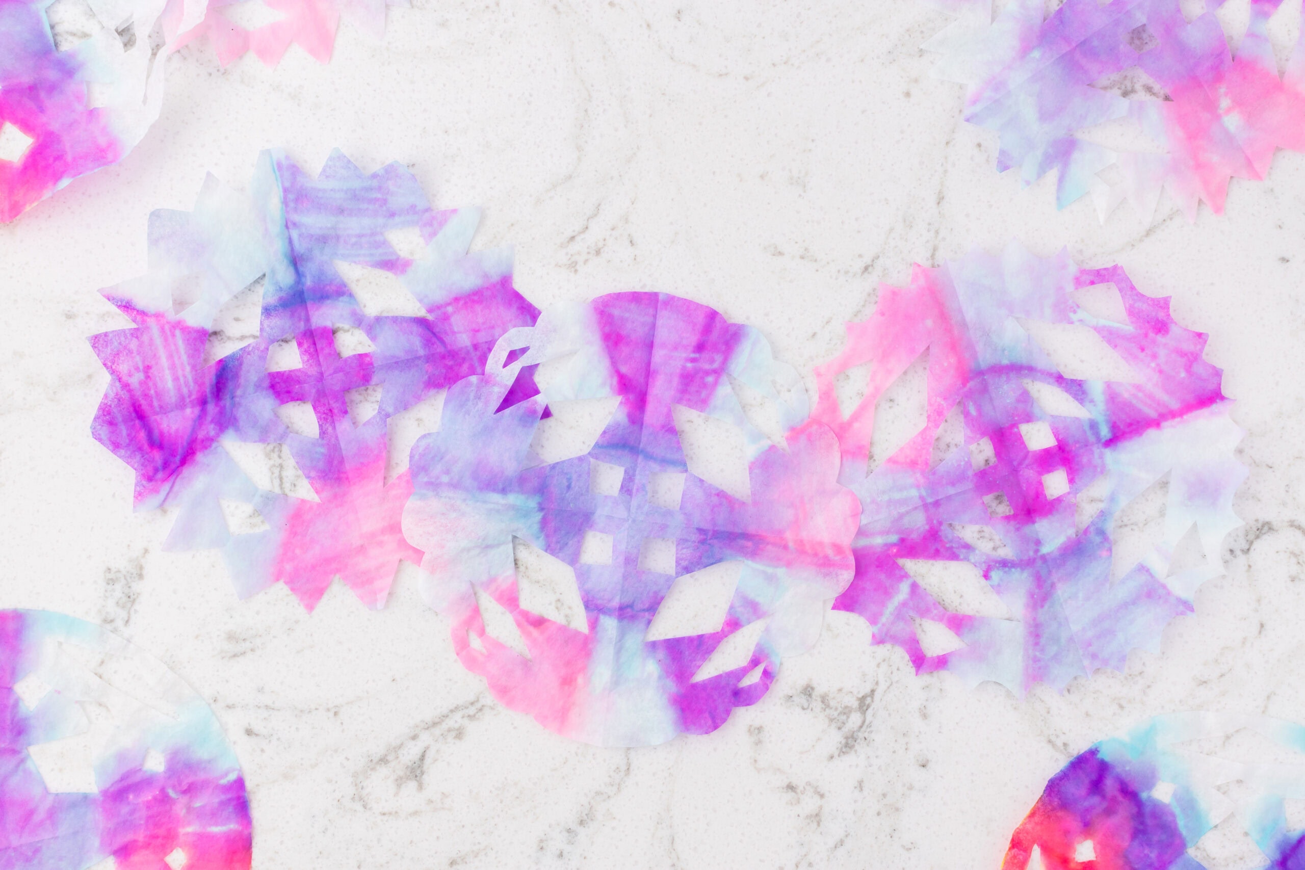 Several pastel tie-dye coffee filter snowflakes in pink, purple, and blue shades laid flat on a white marble surface.