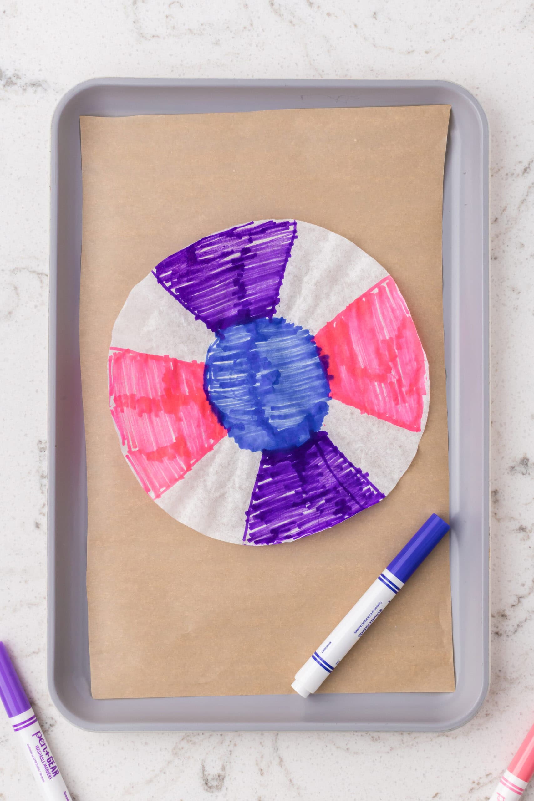 A coffee filter colored with purple, pink, and blue marker sections lying on parchment paper on a tray, with markers nearby.