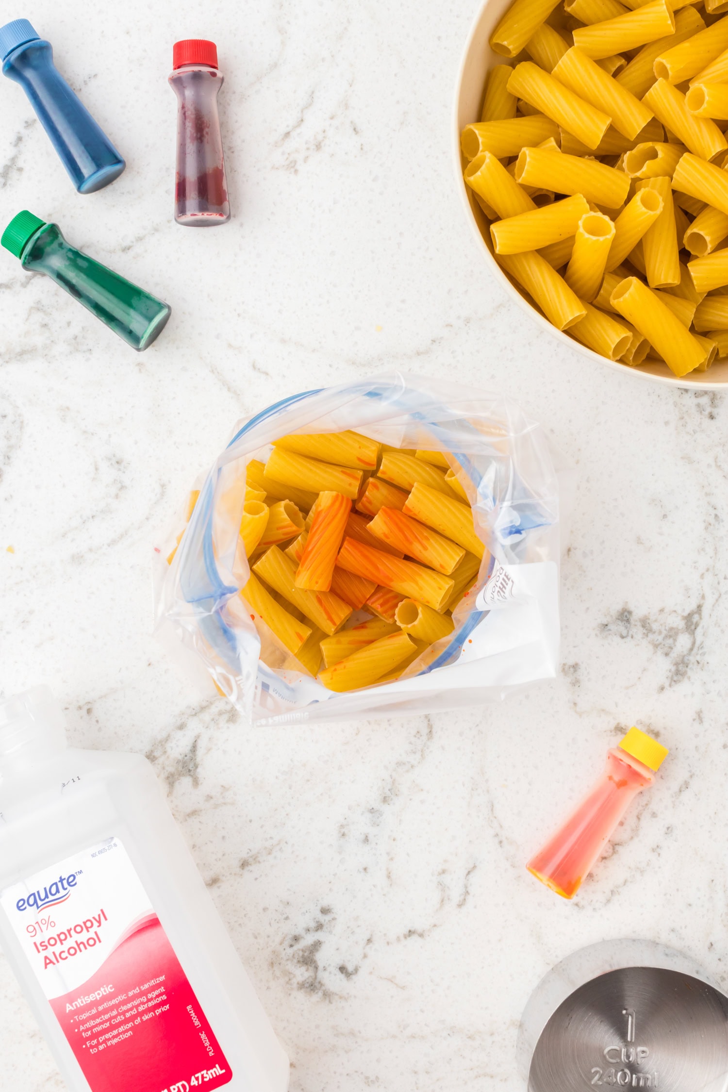 Dry pasta in bowls and plastic bags with food coloring bottles and rubbing alcohol for dyeing pasta crafts.