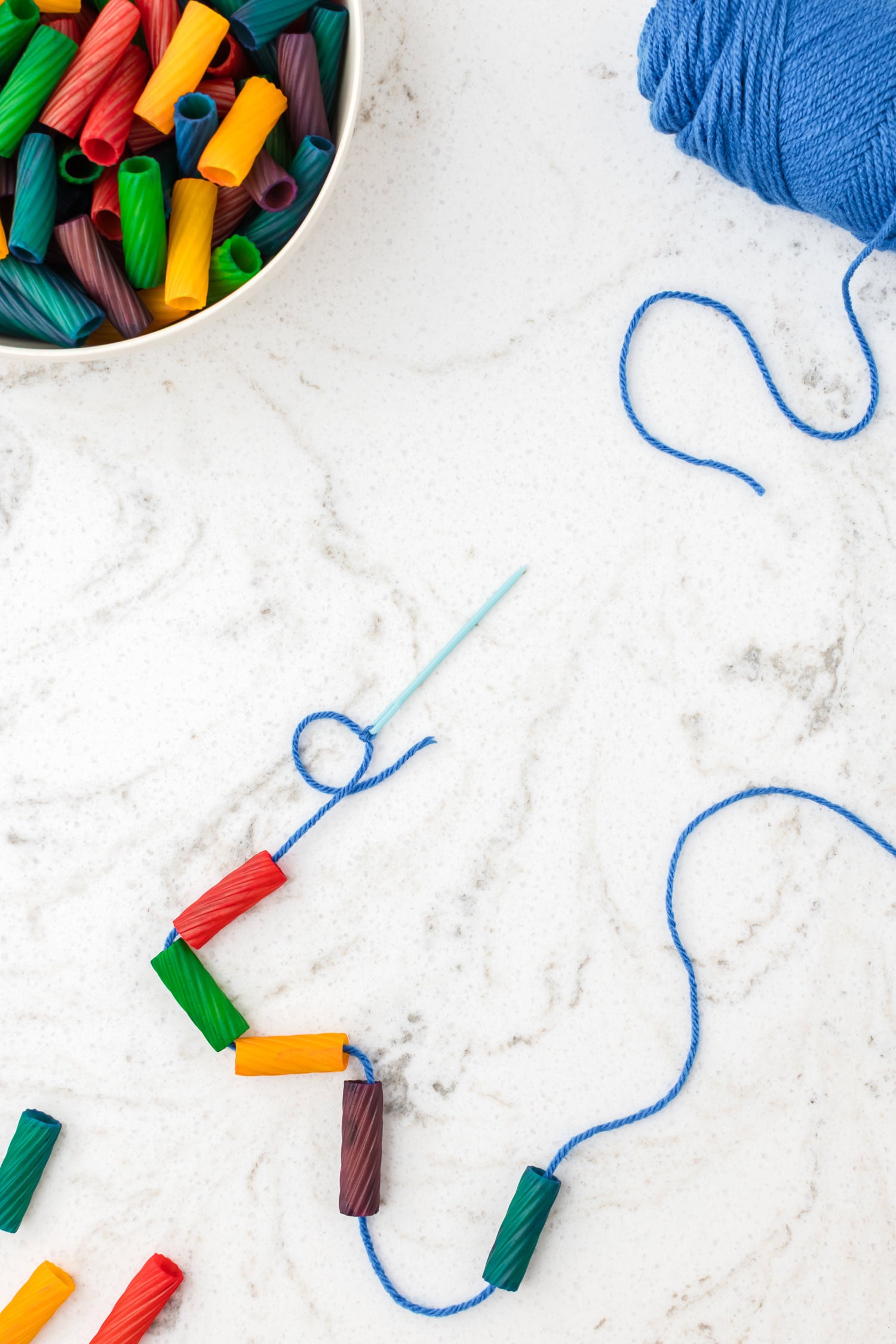 Colored pasta pieces being threaded onto blue yarn with a plastic needle to make a pasta necklace.