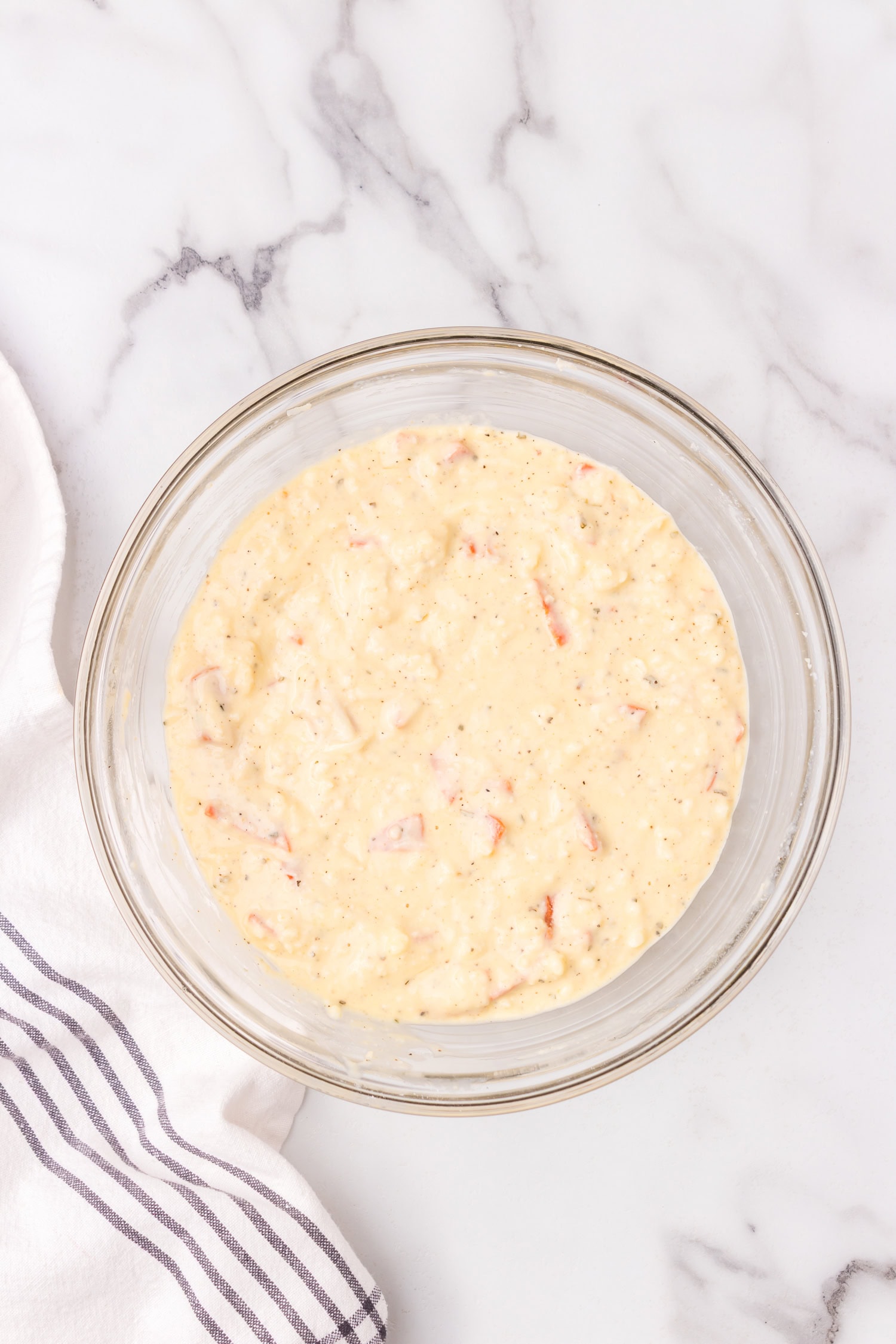 Glass bowl filled with thick pizza muffin batter mixed with shredded cheese, diced pepperoni, and herbs on a marble countertop.
