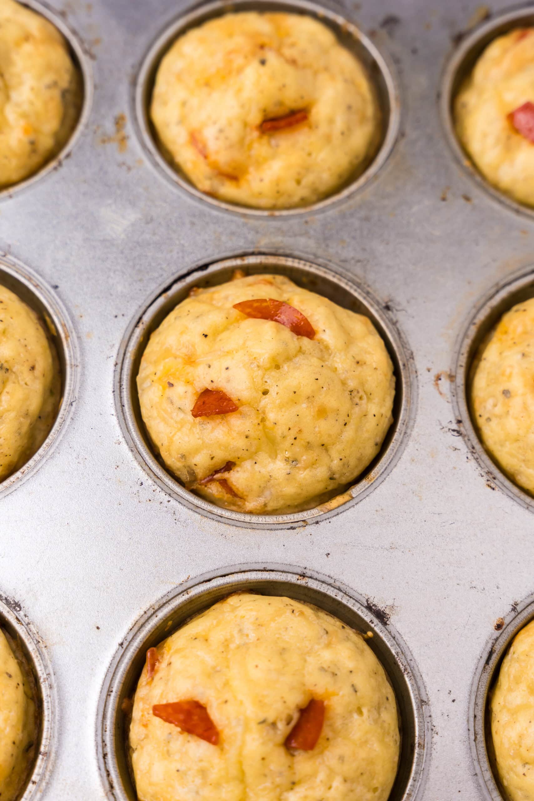 Close-up of baked pizza muffins in a muffin tin, showing lightly browned tops with bits of pepperoni and Italian seasoning.