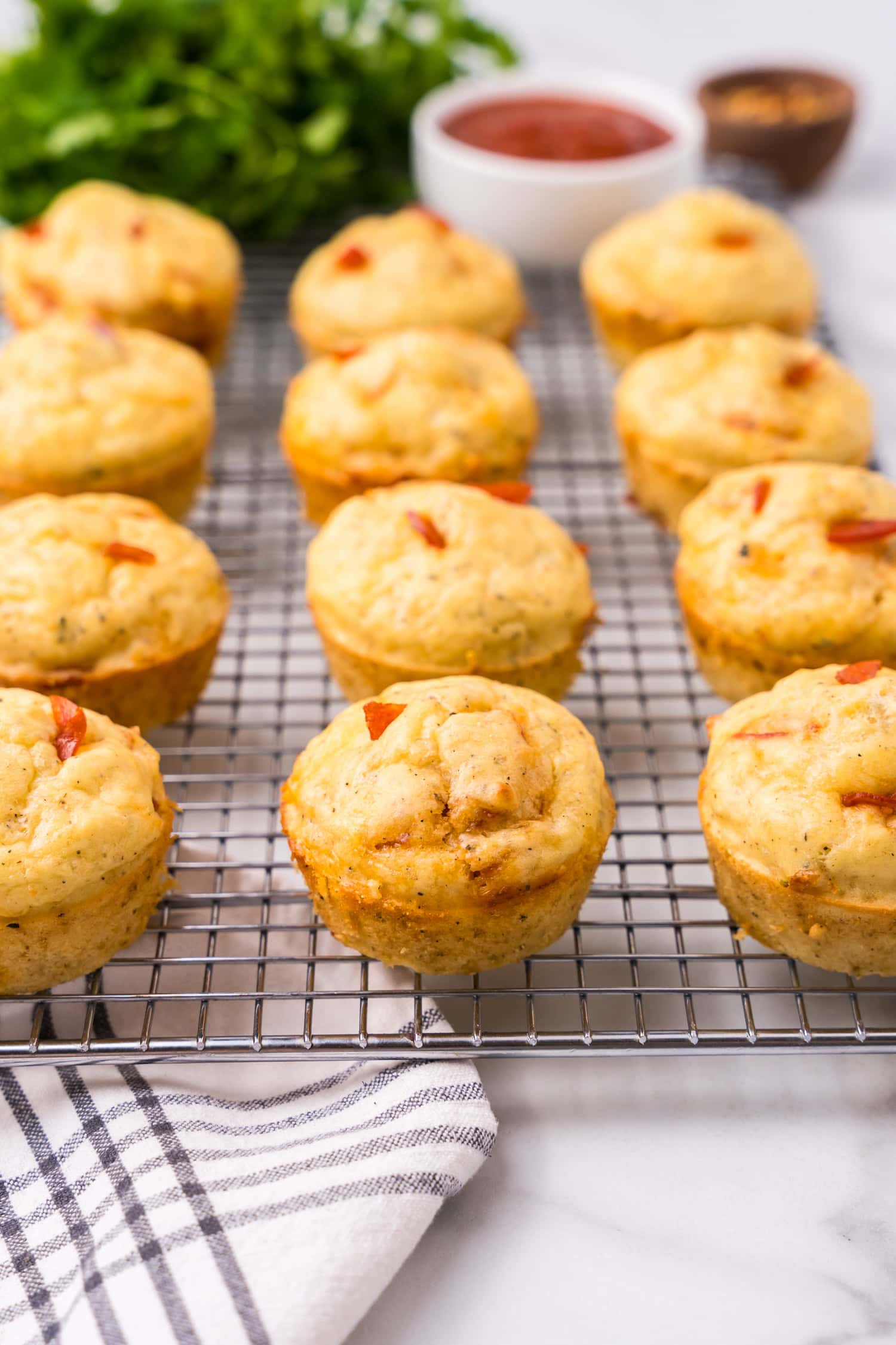 Pizza muffins cooling on a wire rack with a bowl of marinara sauce and fresh herbs blurred in the background.