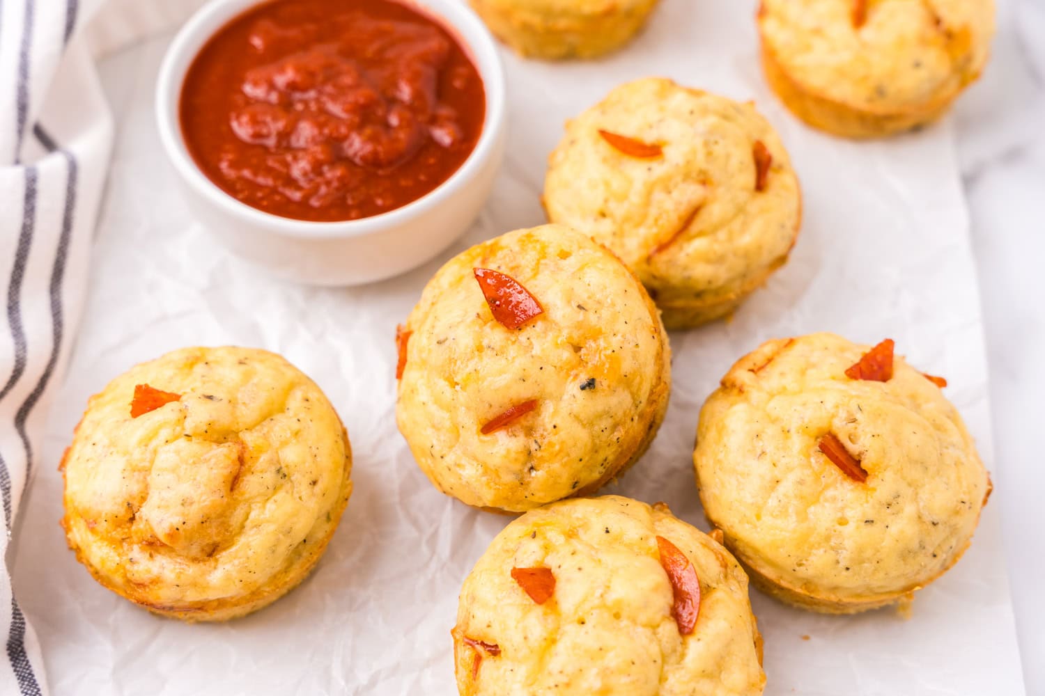 Several golden pizza muffins arranged on parchment paper beside a small bowl of marinara dipping sauce.