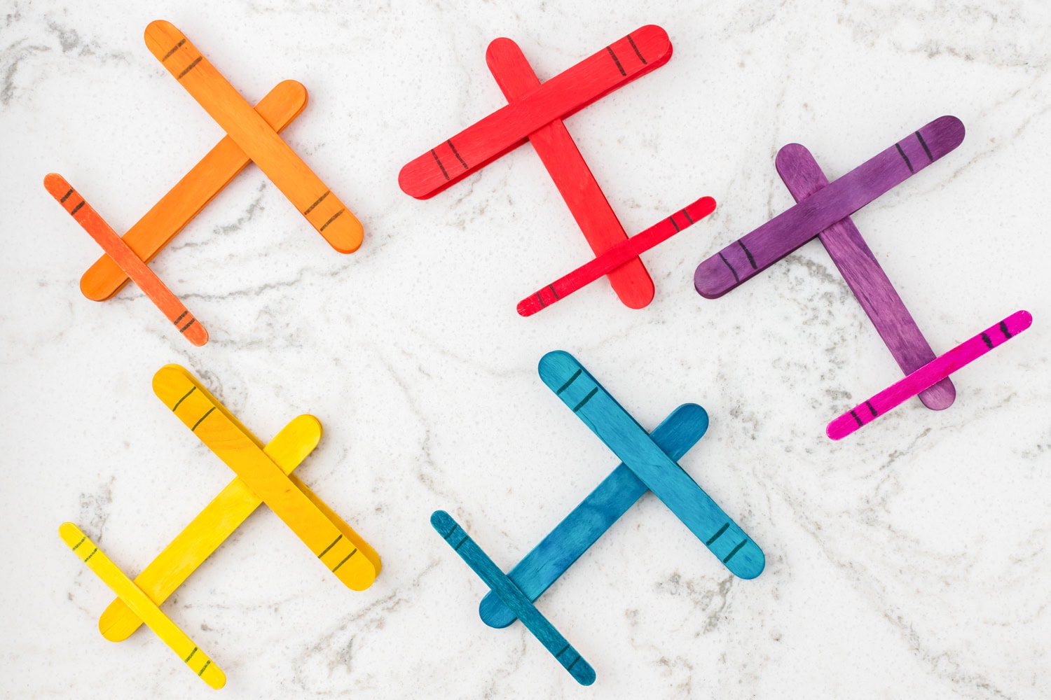 Colorful popsicle stick airplanes in orange, red, purple, yellow, blue, and pink arranged on a marble background.