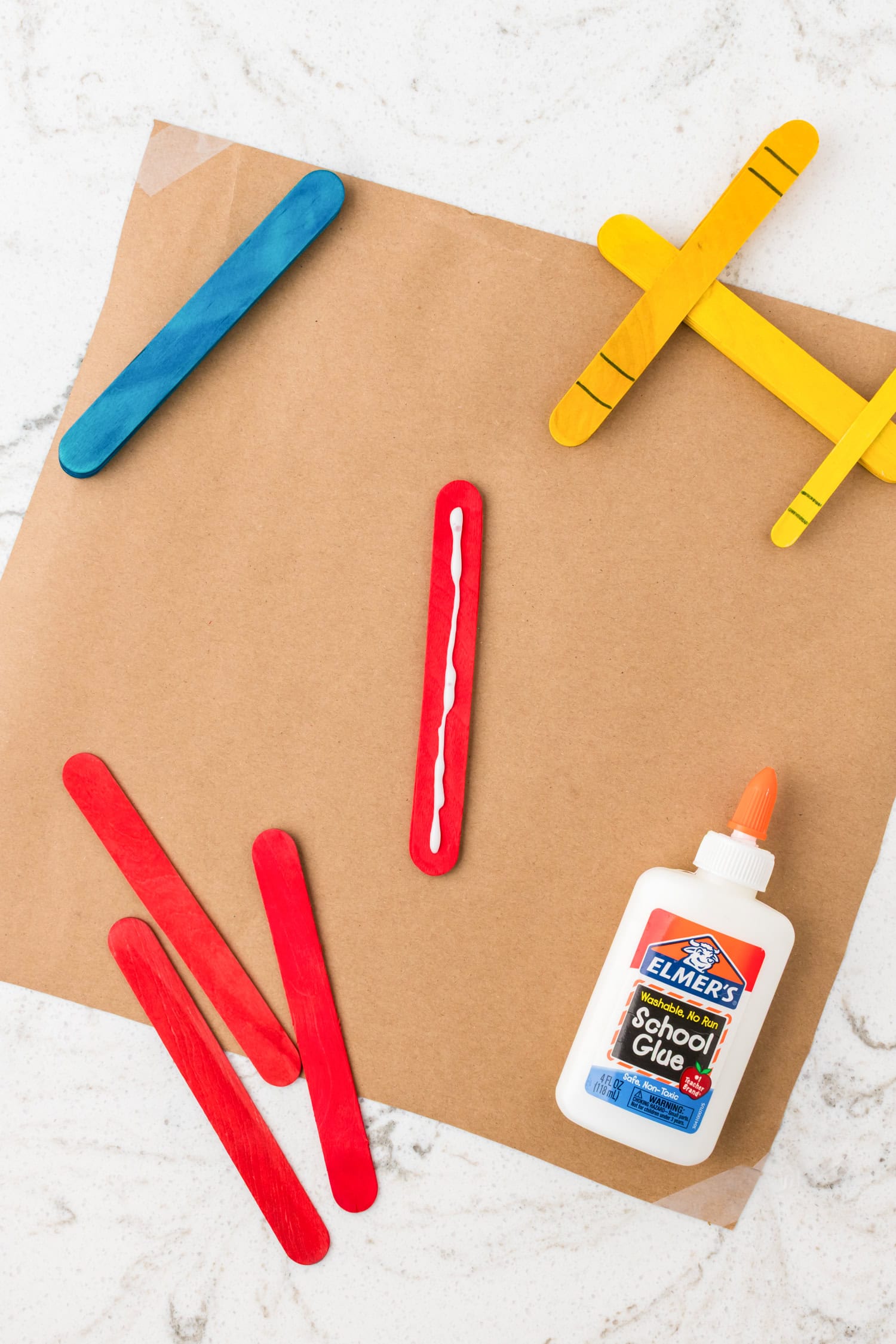 Red and yellow painted popsicle stick airplanes on brown craft paper, with a bottle of school glue on a white marble surface.