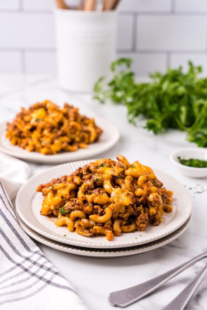 Plate of cheesy beefaroni with ground beef and elbow macaroni topped with melted cheddar, with a second plate blurred in the background on a marble countertop.