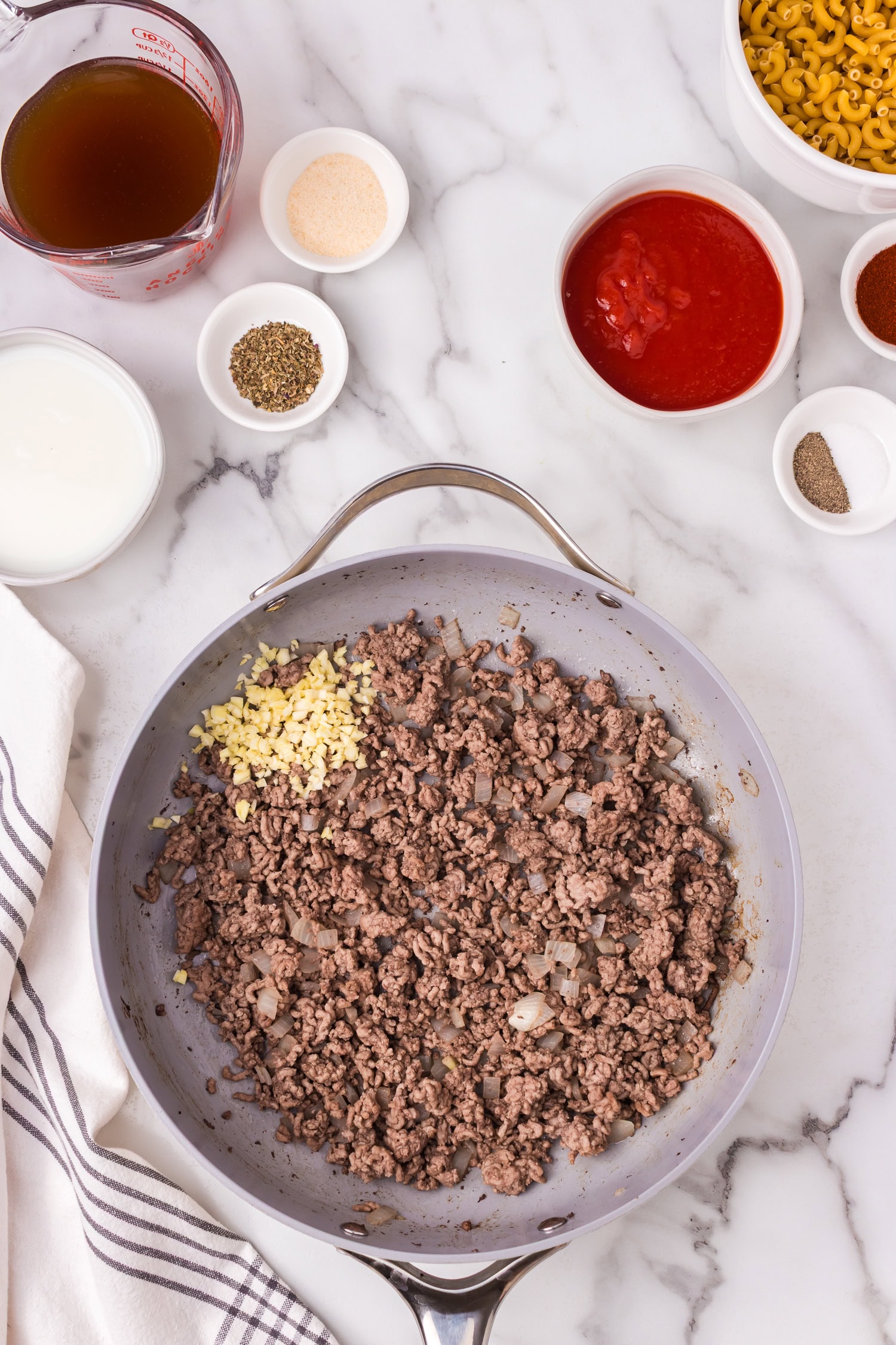 Skillet with browned ground beef, onions, and minced garlic, surrounded by ingredients including broth, tomato sauce, milk, spices, and dry elbow macaroni.