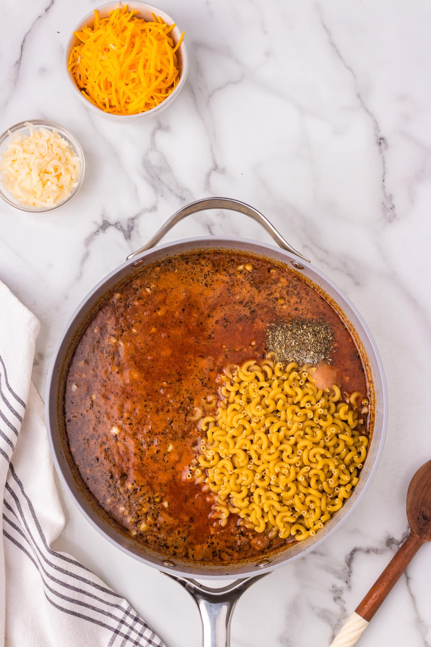 Skillet filled with broth and tomato sauce with uncooked elbow macaroni and seasonings added, cheese bowls nearby on a marble surface.