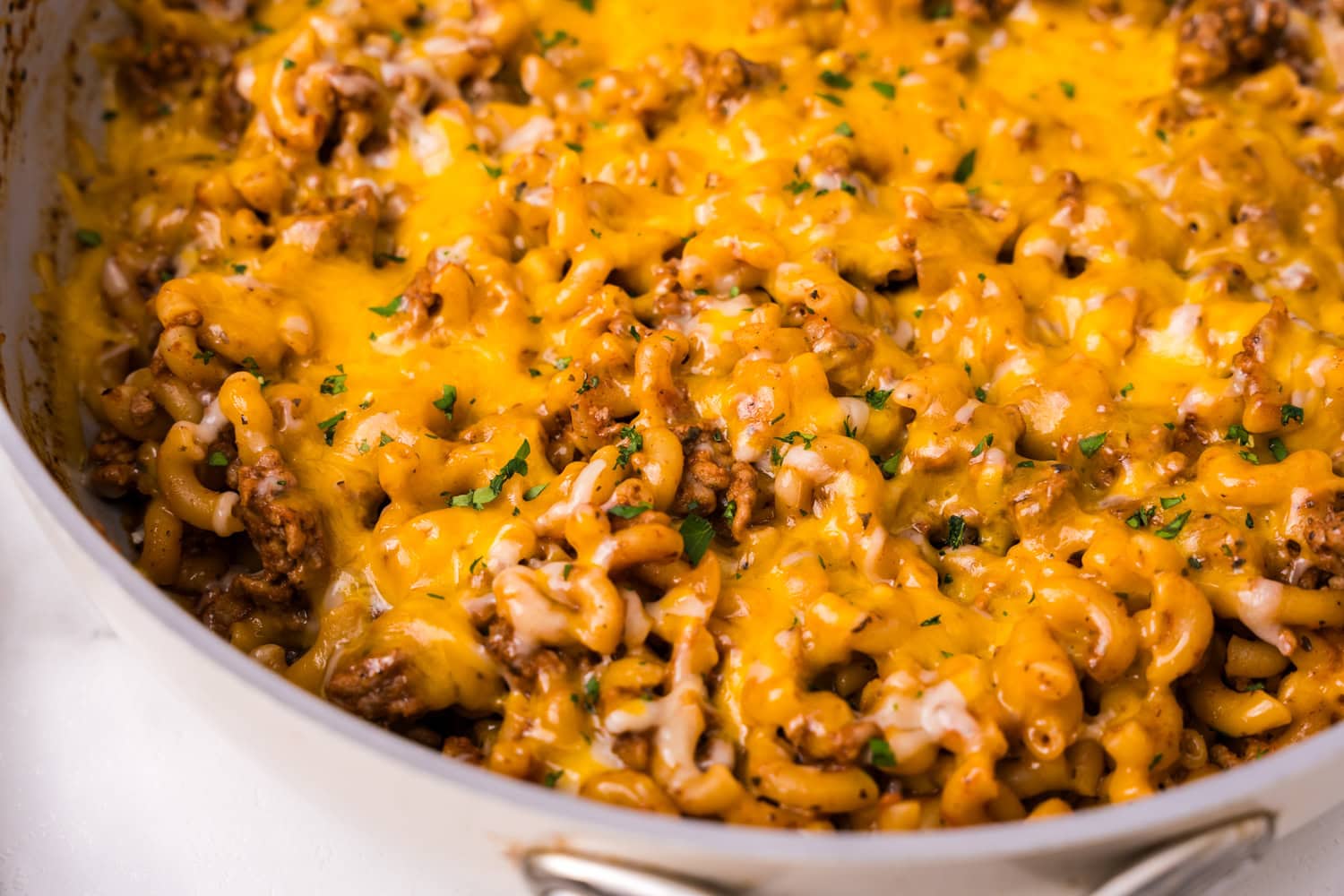 Close-up of skillet filled with cheesy beefaroni, showing melted cheddar over elbow macaroni and seasoned ground beef, garnished with herbs.