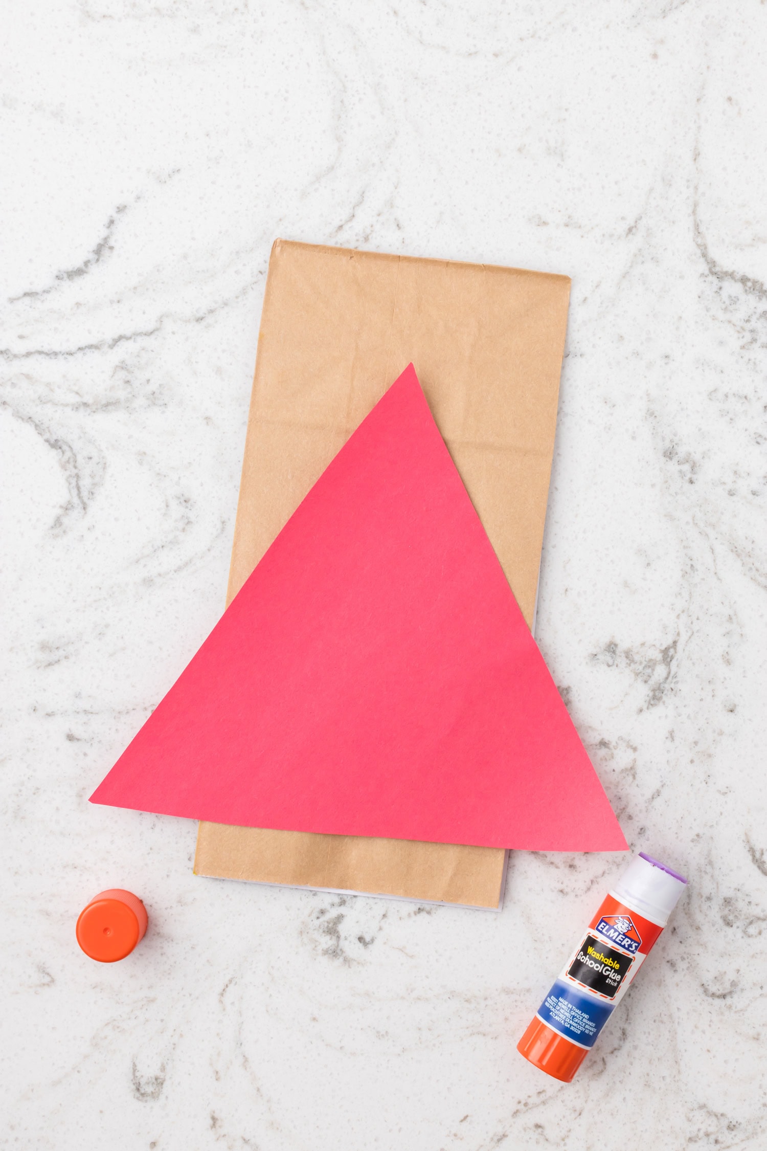 Triangle of red paper glued onto a brown paper bag to start a superhero craft, with a glue stick beside it.