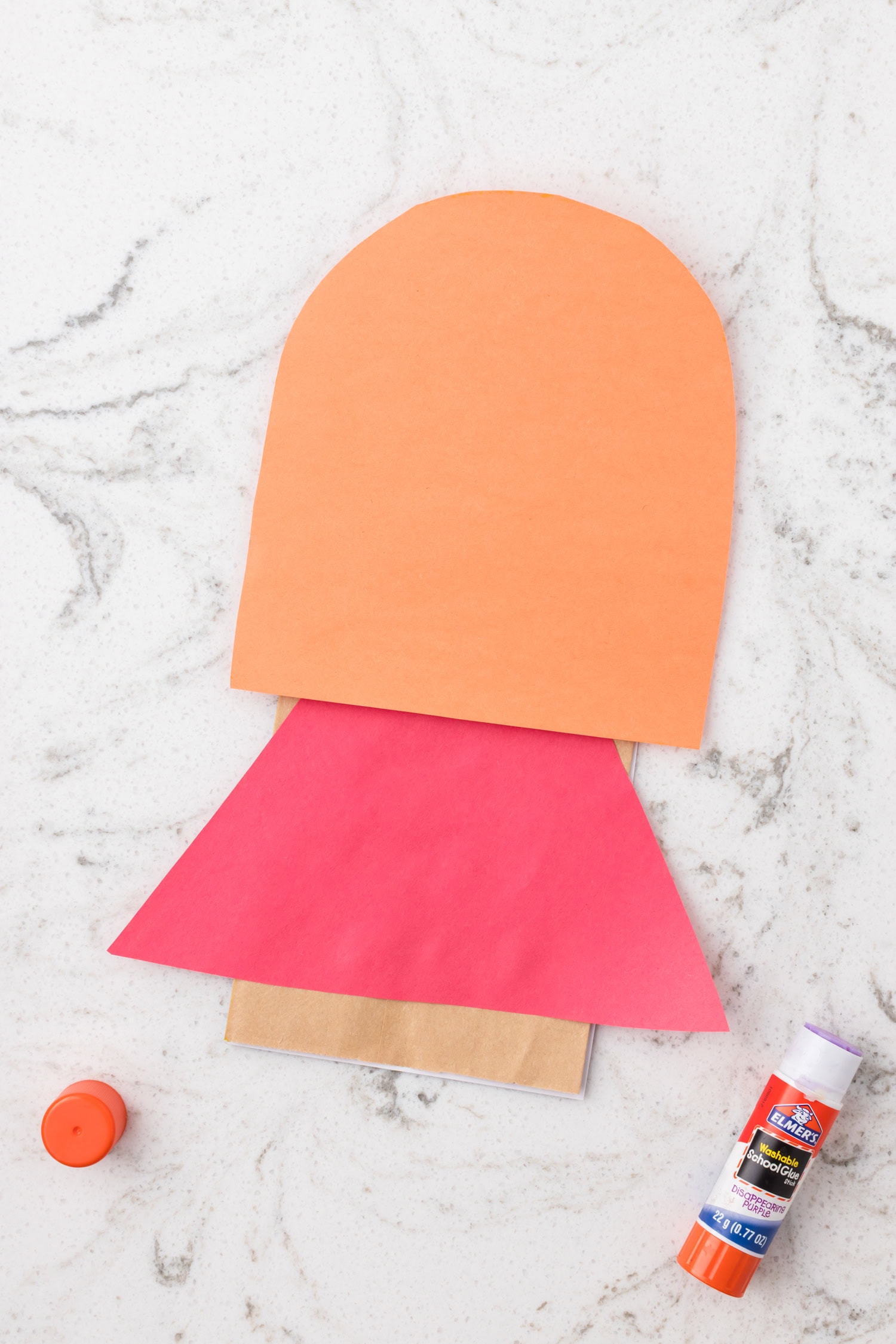 Orange rounded paper piece added above the red triangle on a paper bag to form the head and cape base for a superhero craft.