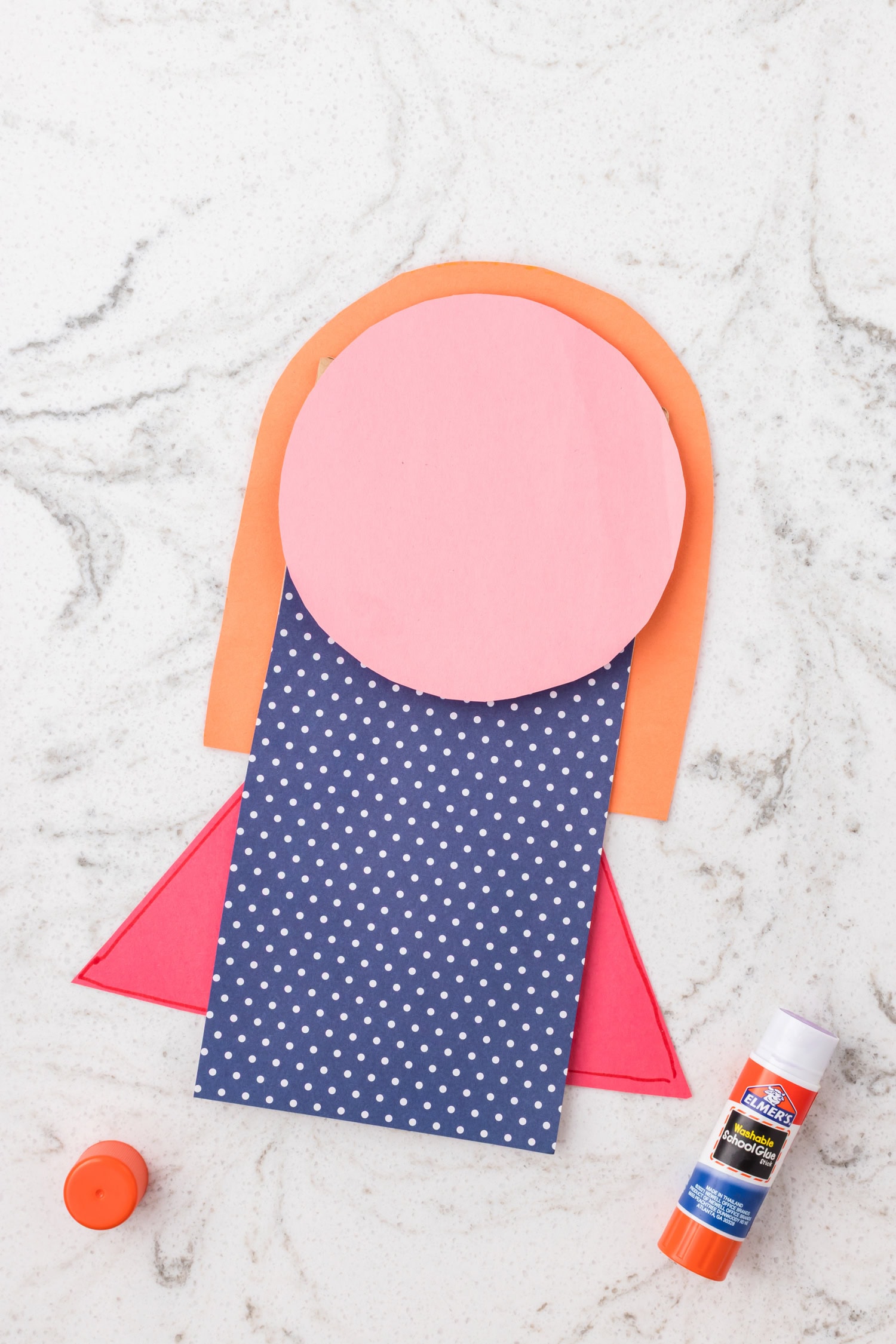 Paper superhero craft in progress with a pink circle face, orange hair, and a blue polka dot shirt on a paper bag.