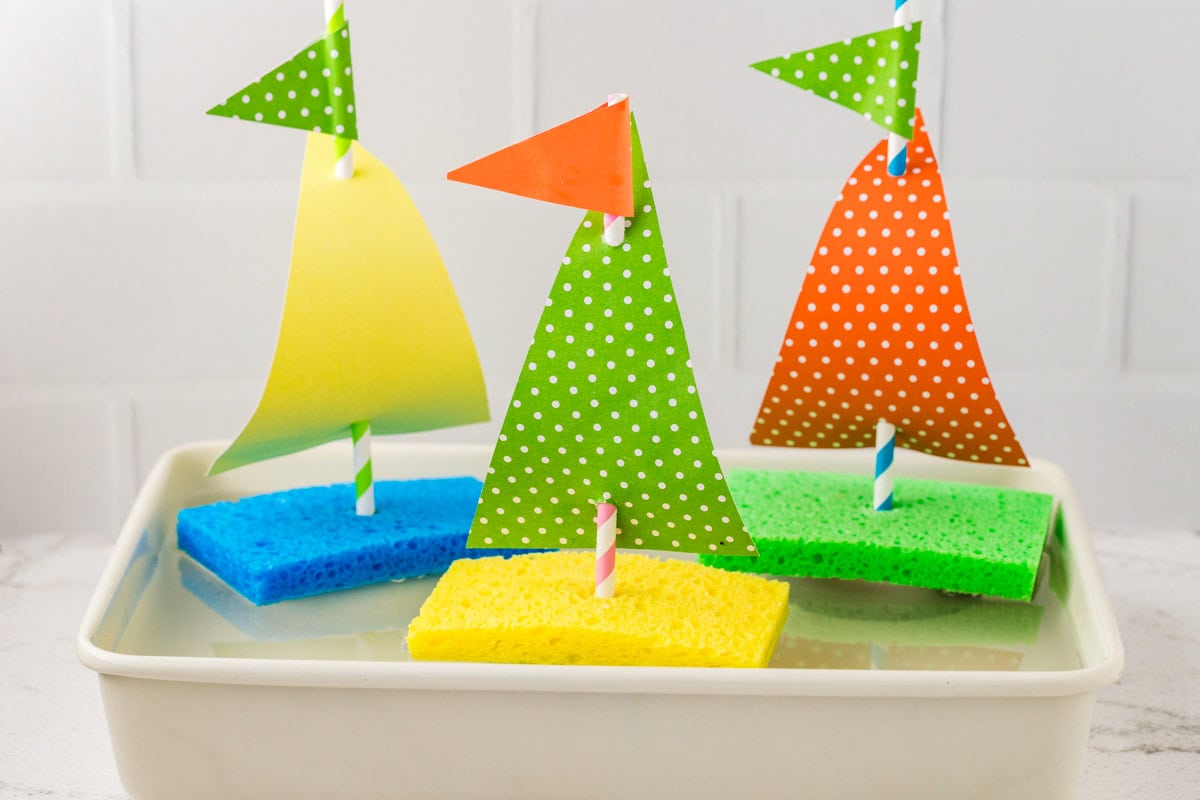 Three colorful sponge sailboats floating in a shallow white bin of water, each made with sponges, paper sails, and striped paper straws.