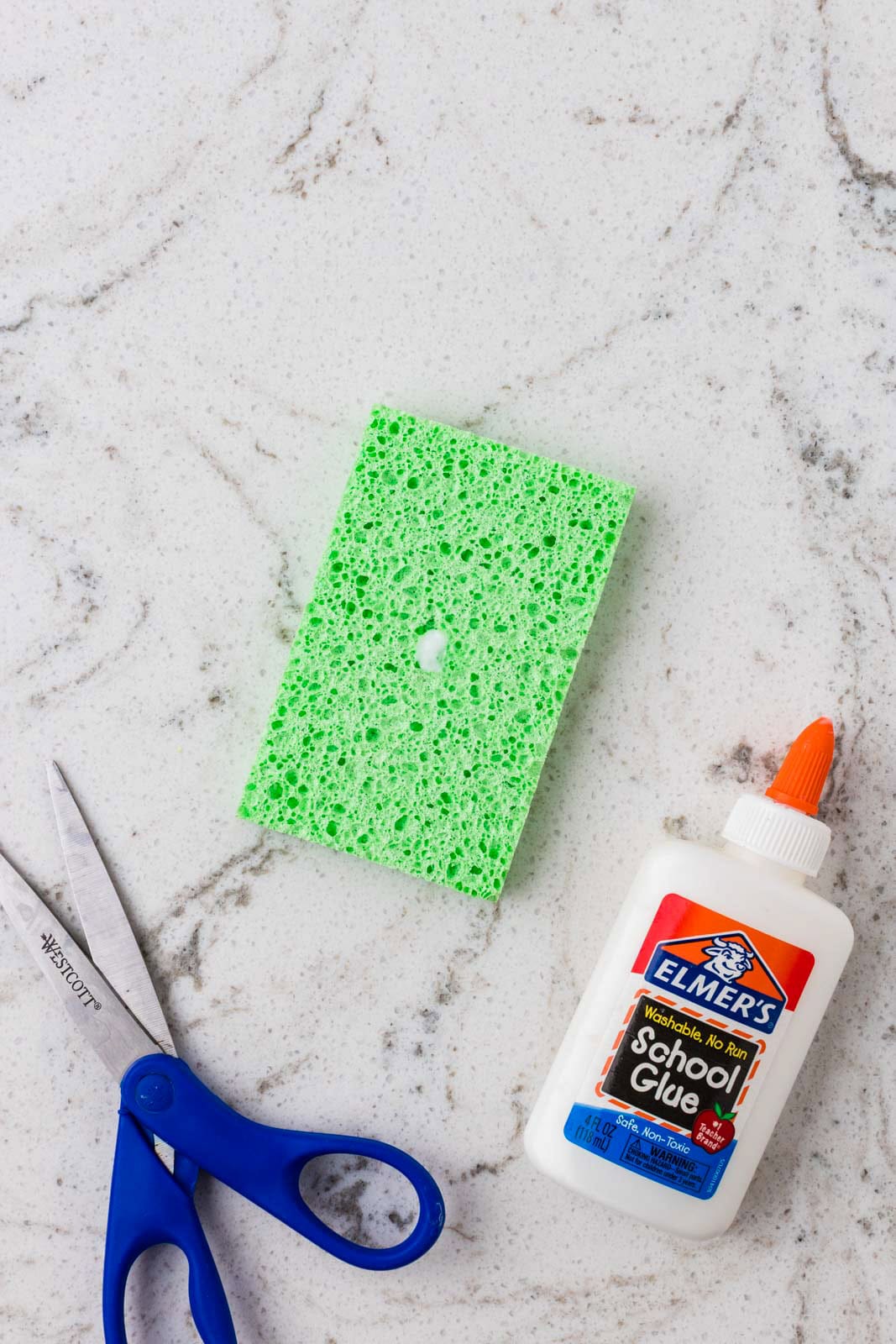 Green sponge rectangle with a hole in the center beside blue-handled scissors and a bottle of school glue on a marble countertop.