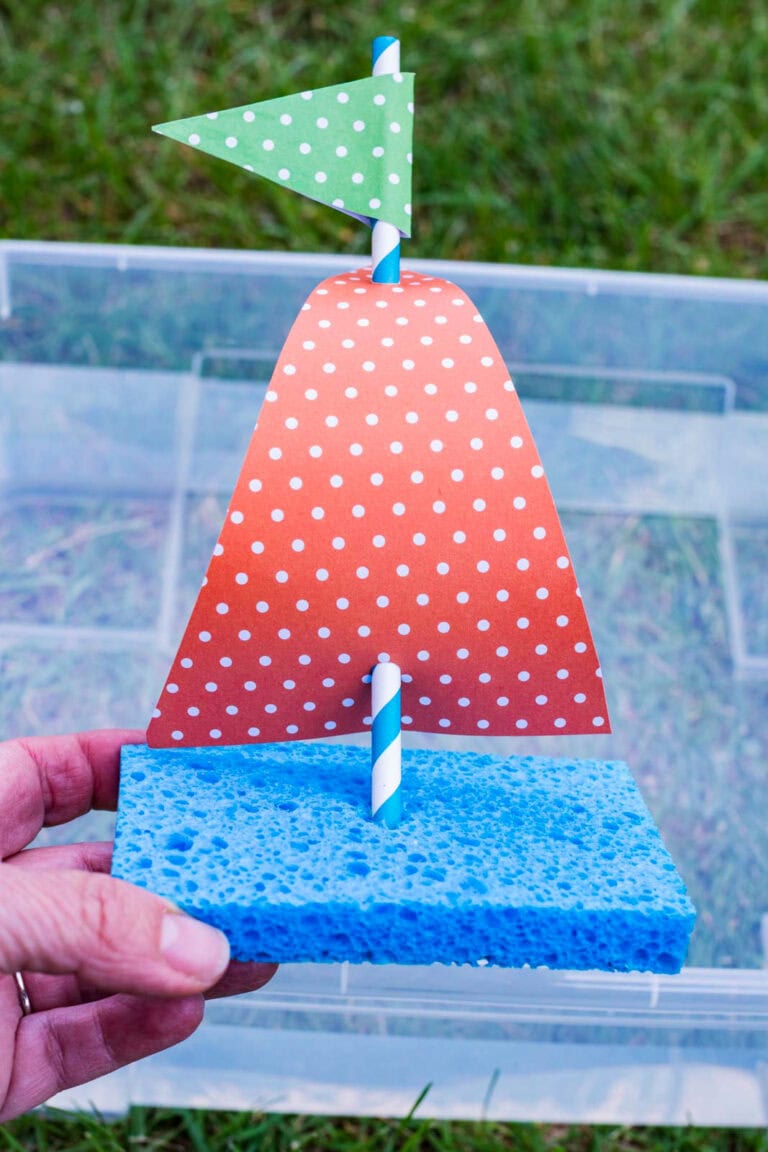Hand holding a completed sponge sailboat craft with a blue sponge base, striped straw mast, and polka dot paper sails over a clear plastic bin outdoors.