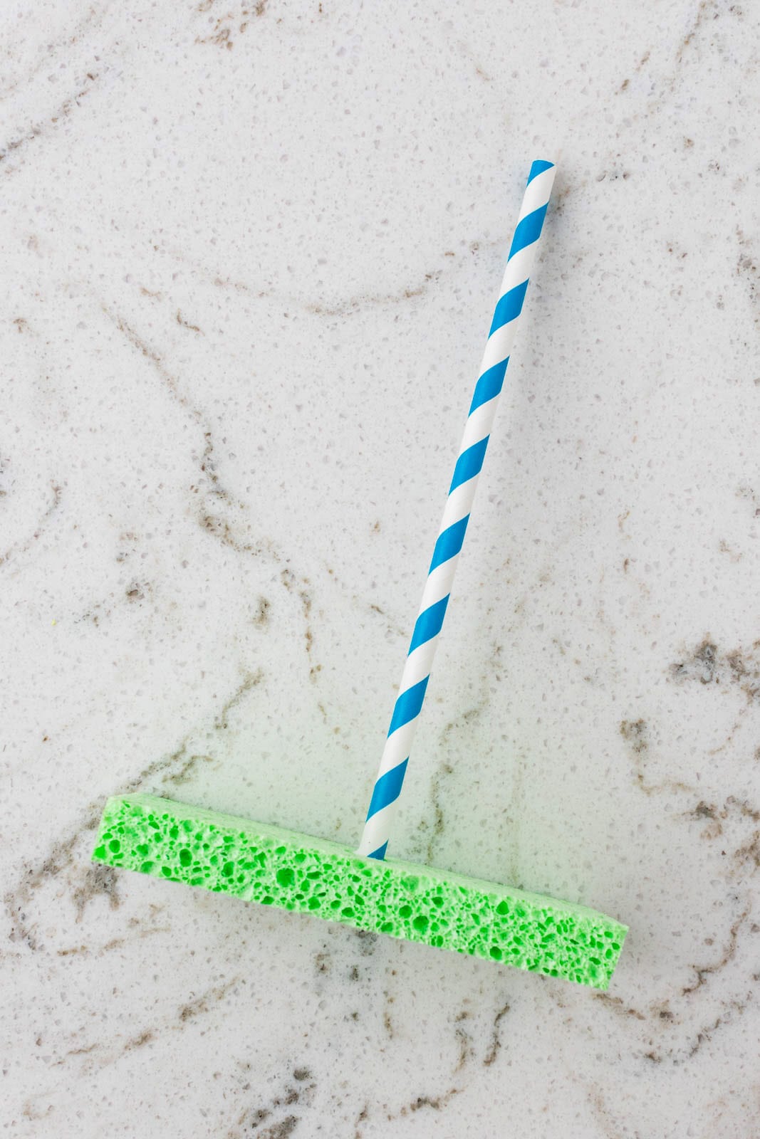 Blue-and-white striped paper straw inserted through a green sponge base to form the mast of a sponge sailboat craft.
